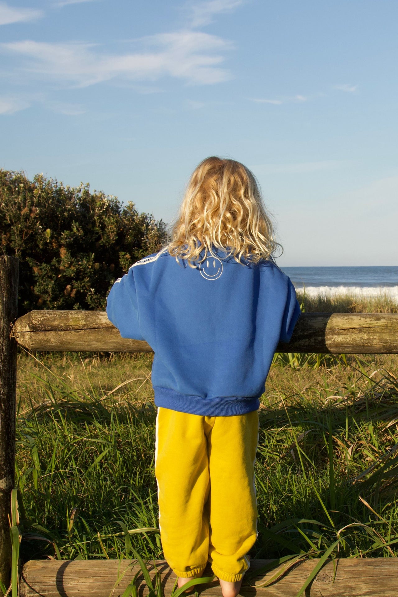 little boy wearing a blue oversized sweater by cracker day club showing the back embroidery while looking at the waves at the beach in australia