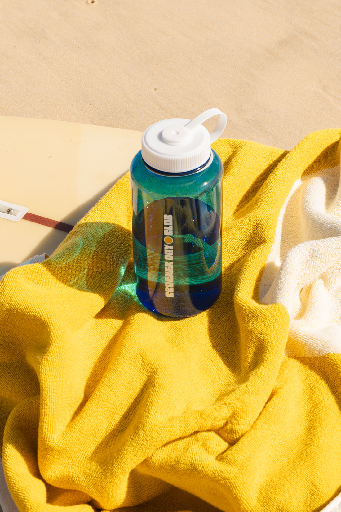 a see through blue water bottle by cracker day club sitting on a luxe towel and longboard surfboard