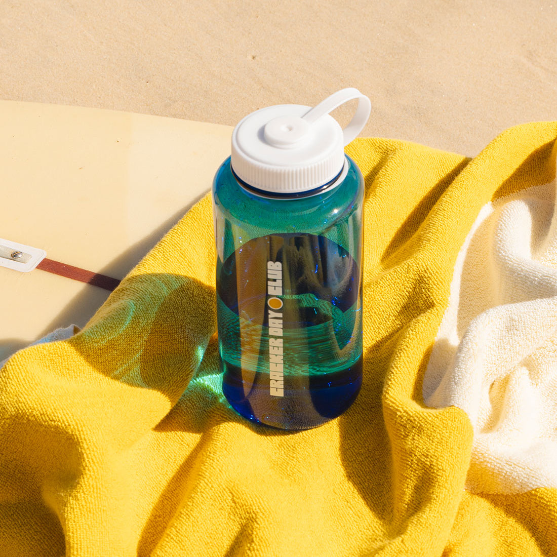 a see through blue water bottle by cracker day club sitting on a luxe towel and longboard surfboard