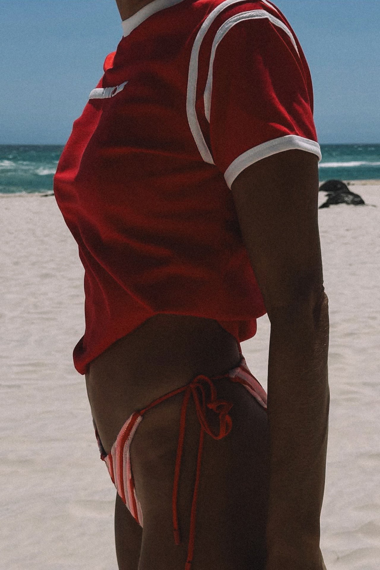 Person wearing a red cracker day club baby tee and bikini bottoms on a beach