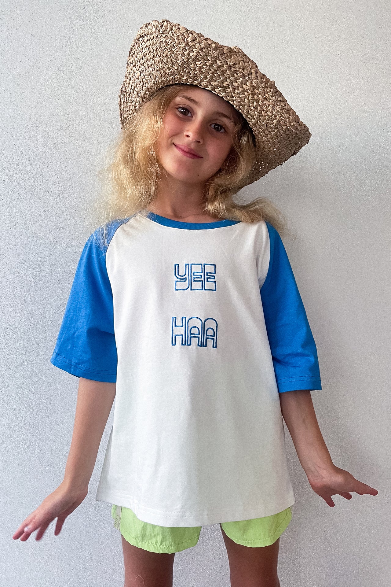 Child wearing a white t-shirt with blue text and blue boardshorts, sitting on a white surface.