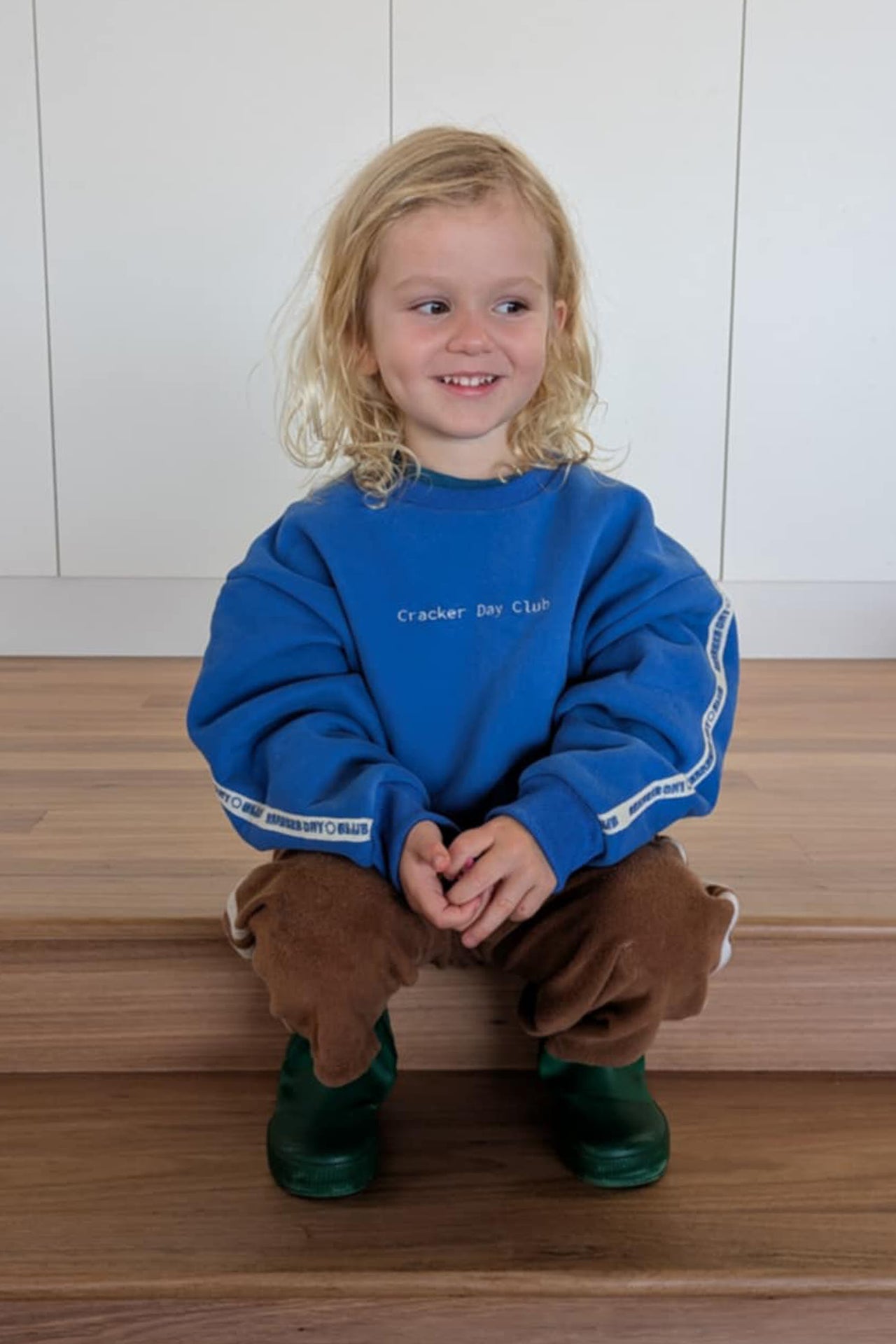 little boy wearing a blue oversized sweater by cracker day club showing the back embroidery while looking at the waves at the beach in australia