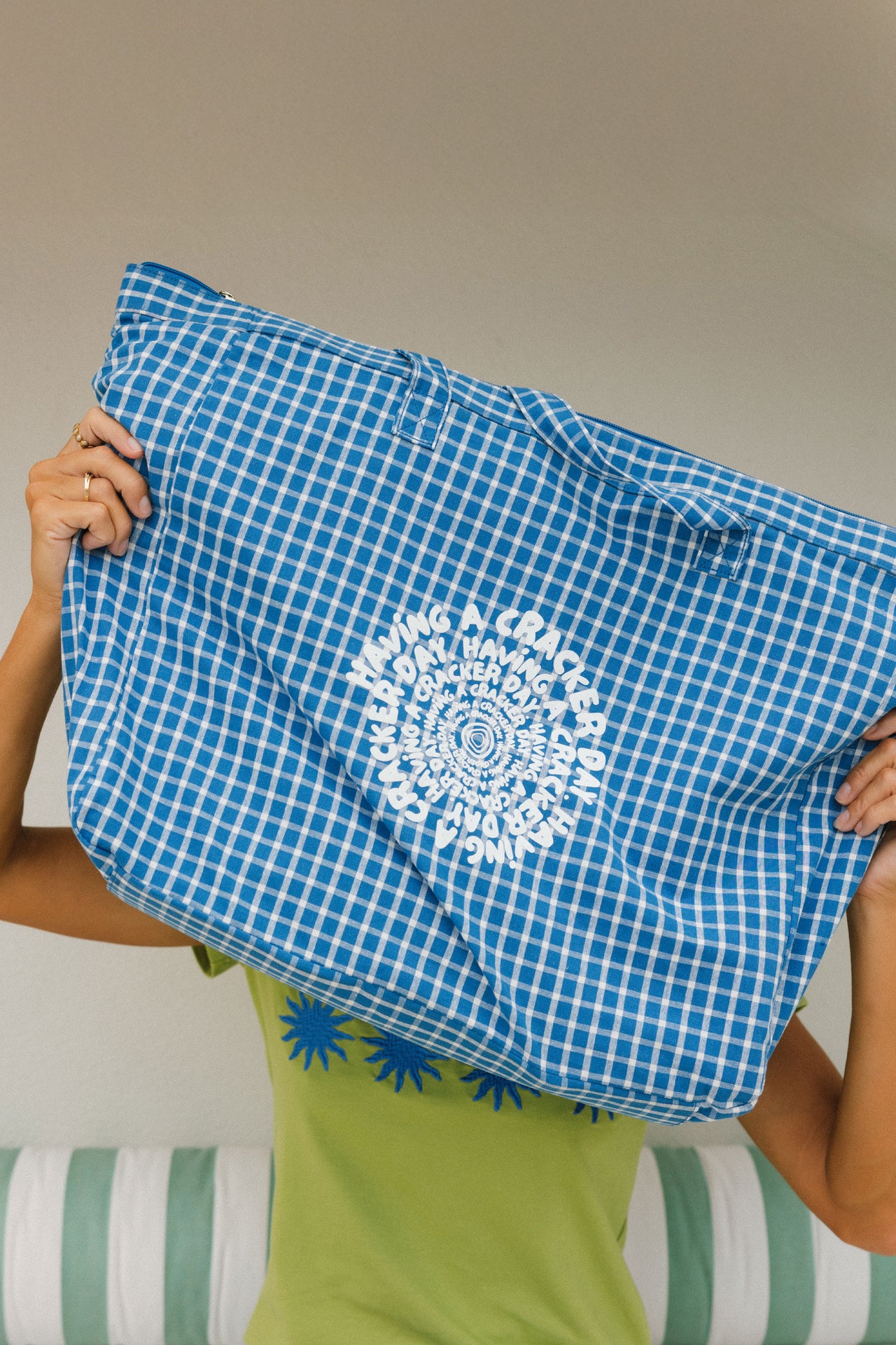 Person holding a blue checkered beach bag with a white cracker day logo design against a neutral background