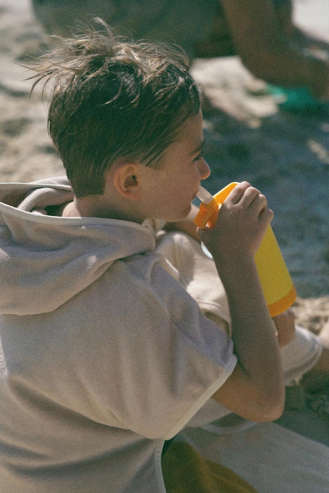 young boy sipping from a drink bottle while sitting on the beach in a retro hoodie towel to keep warm and sun safe