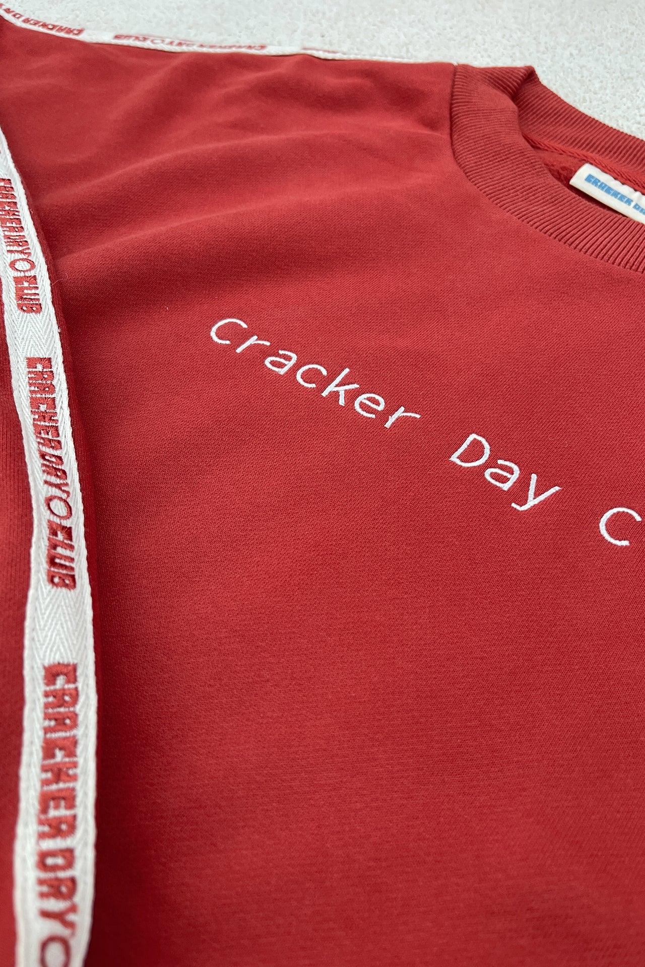 Woman in a cracker day club red sweatshirt sitting on sand with a large orange flower.