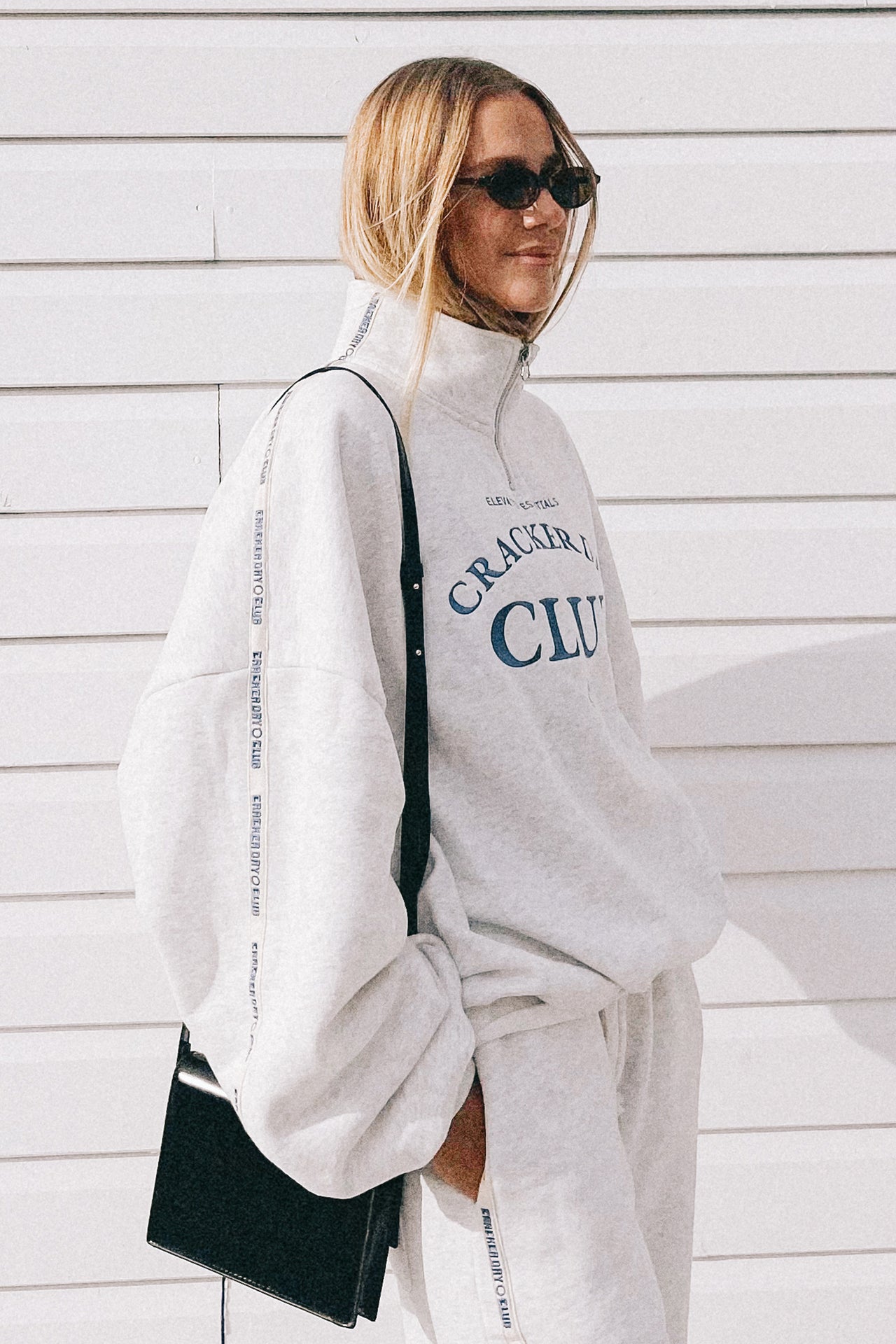 Person wearing a white sweatshirt with text and pants, holding a black bag against a light wooden wall.