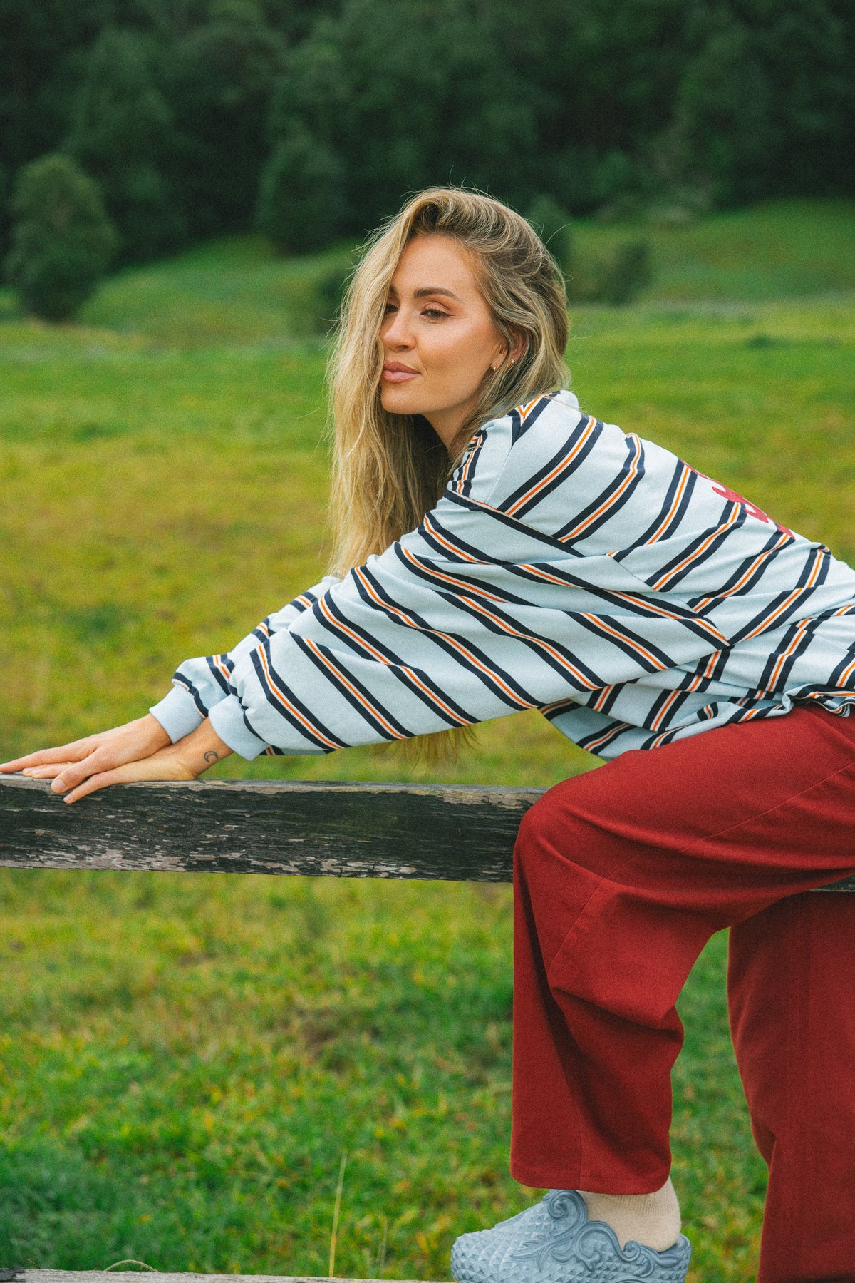 A woman in a striped long-sleeve top and red pants sitting on a wooden fence in a grassy field.