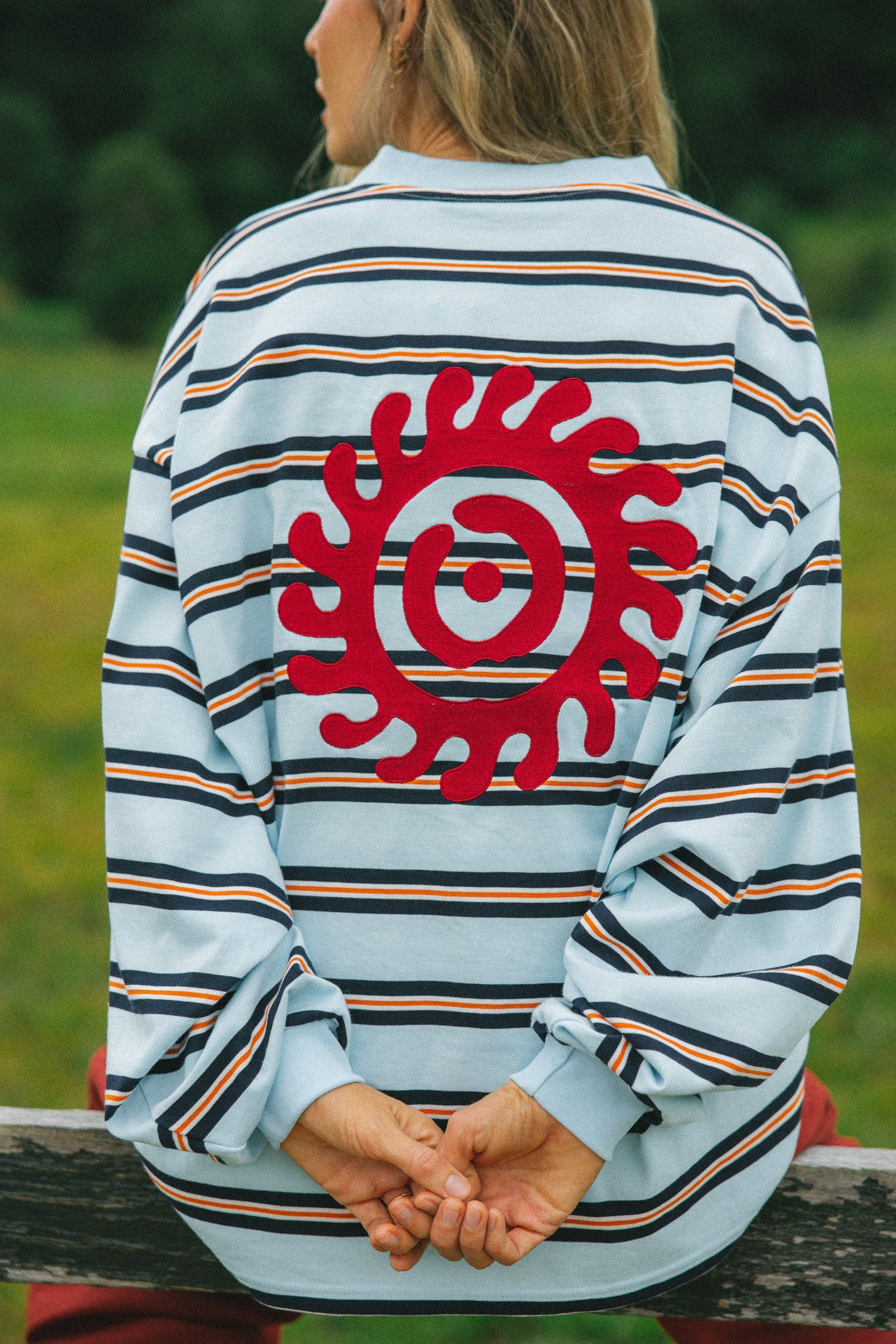 A person viewed from behind wearing a light blue long-sleeve striped shirt with thin navy, orange, and white stripes, featuring a large red circular sun-like graphic on the back. The person is sitting outdoors on a wooden bench with a blurred green natural background.