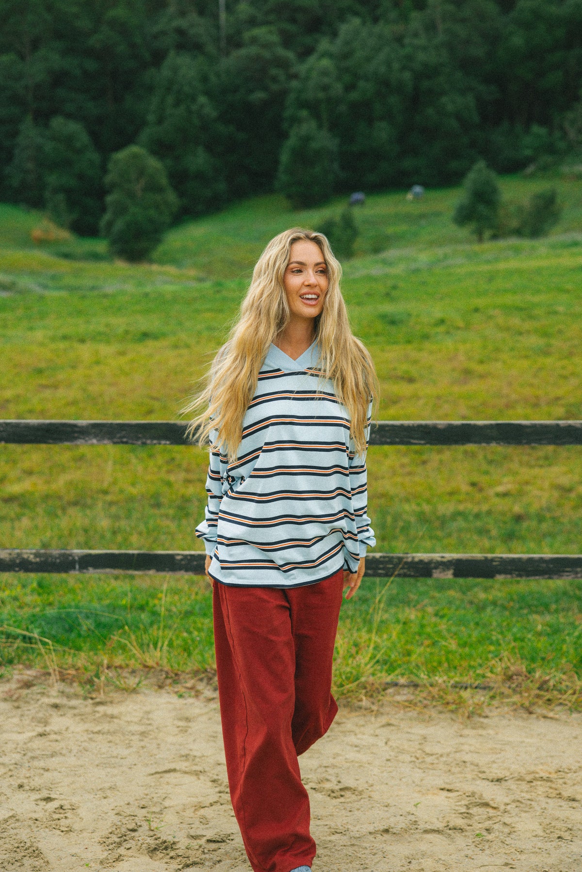 A woman stands outdoors in a grassy field wearing a light blue long-sleeve polo shirt with thin dark horizontal stripes and loose rust-red pants.