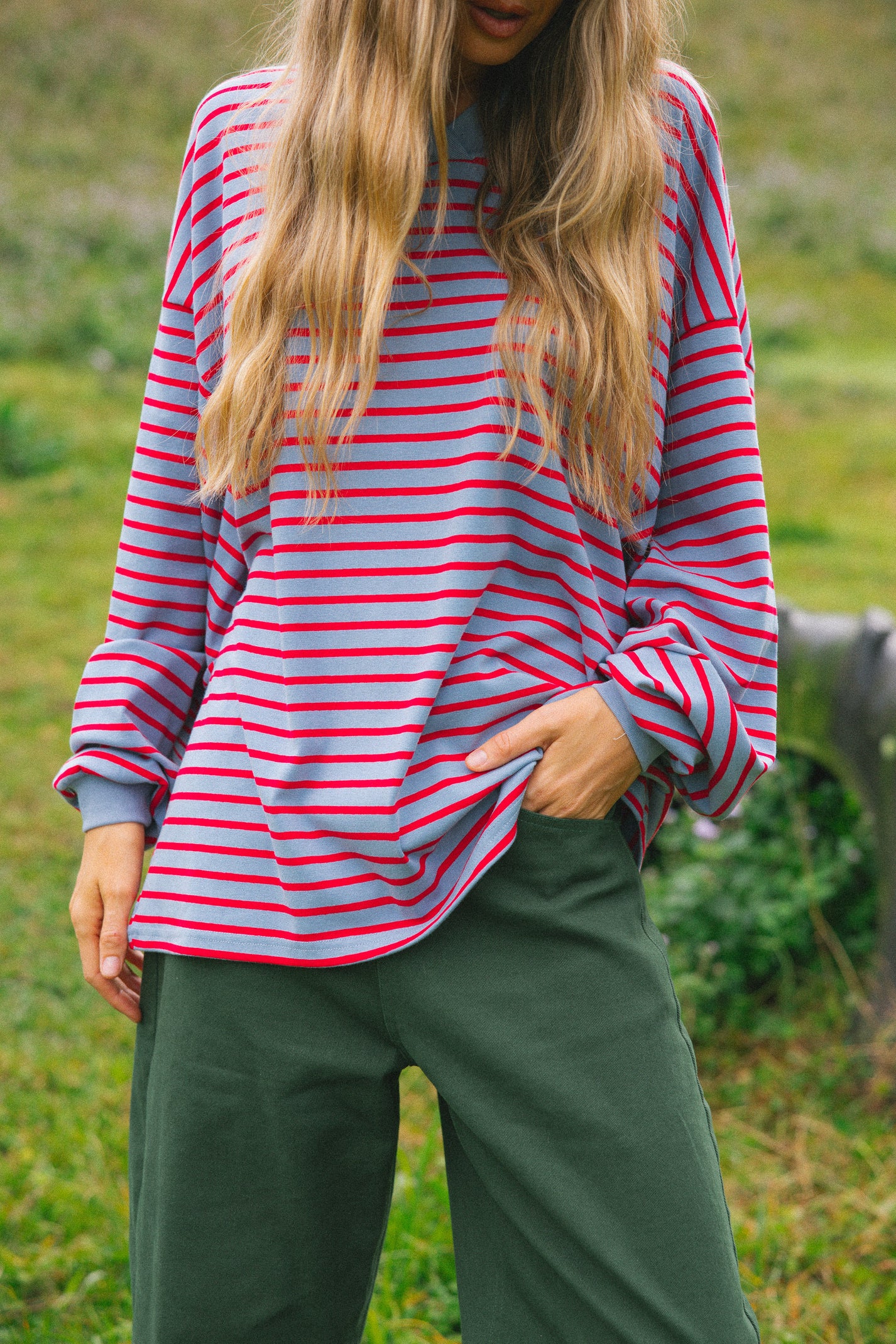 A woman wearing a pink and red striped long-sleeve top with dark green pants, standing outdoors in a grassy field.