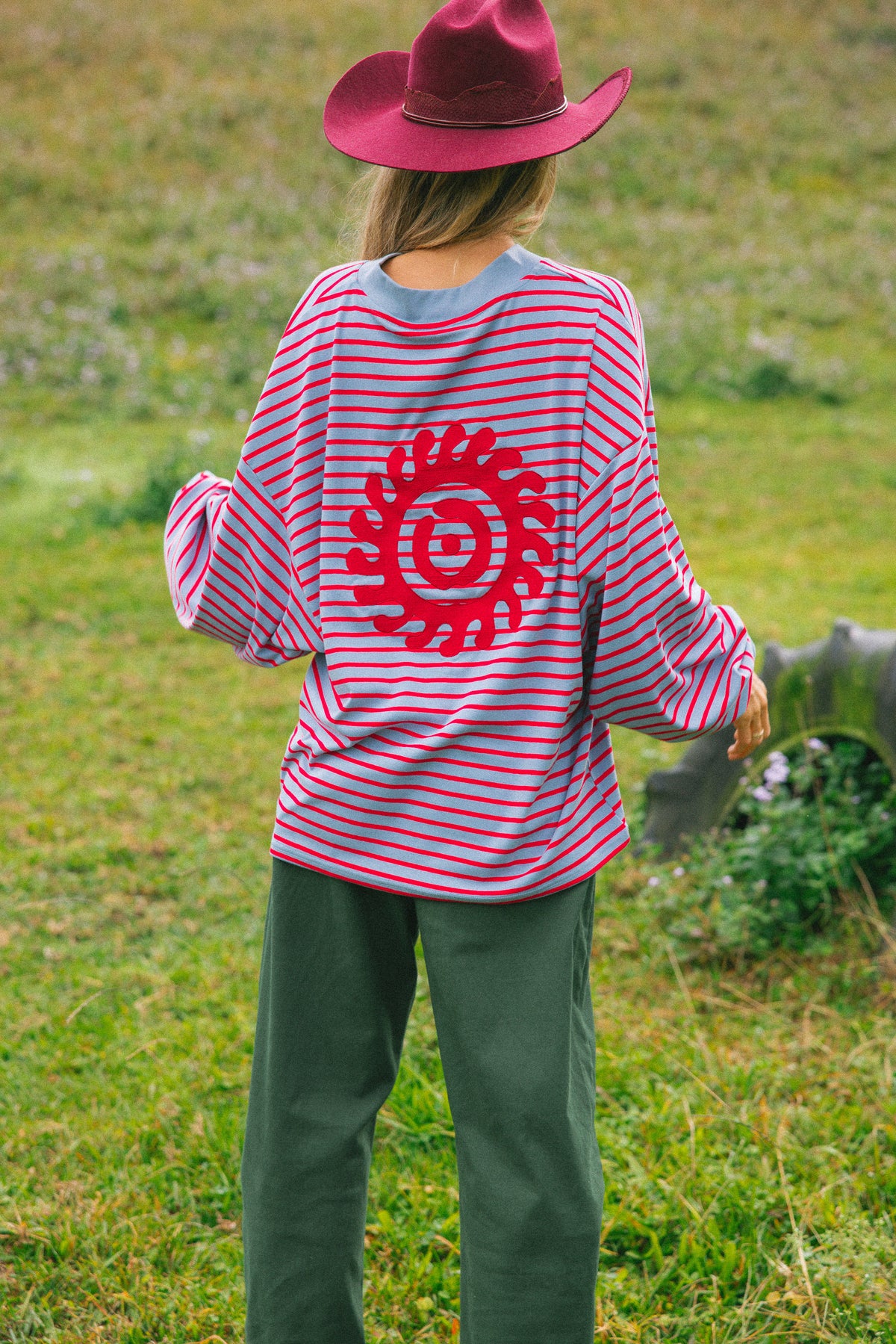 A person standing outdoors in a grassy field, viewed from the back, wearing a pink and red horizontally striped long-sleeve shirt with a large red circular sunburst graphic on the back, dark green pants, and a dark red cowboy hat.