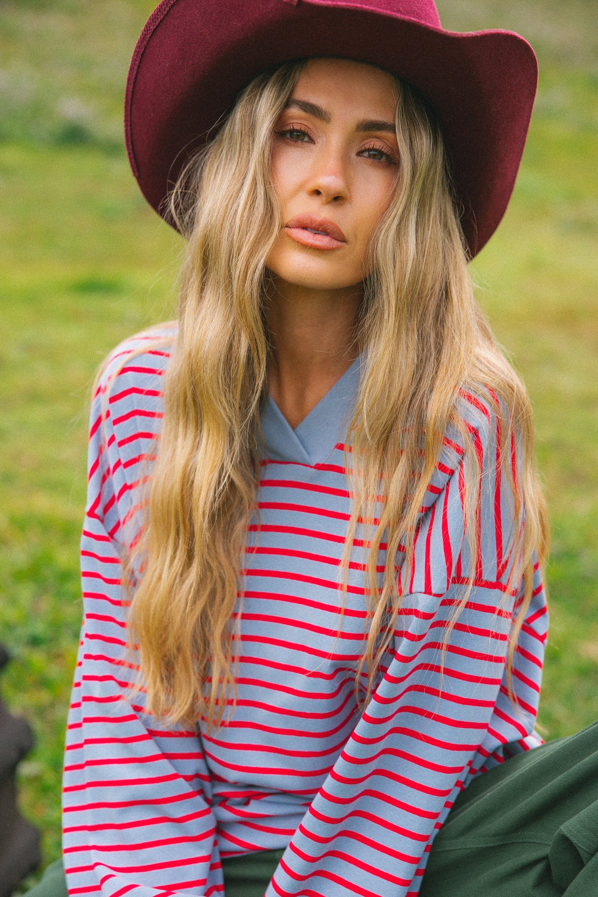 A woman wearing a burgundy wide-brim hat, a red and light blue striped long-sleeve shirt, and dark green pants, sitting outdoors on a grassy field.