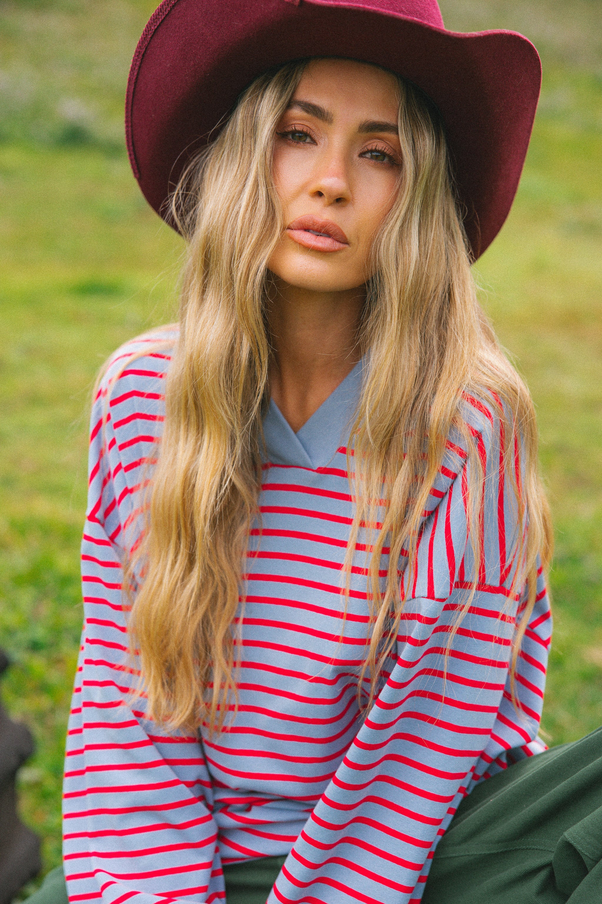 A woman wearing a burgundy wide-brim hat, a red and light blue striped long-sleeve shirt, and dark green pants, sitting outdoors on a grassy field.