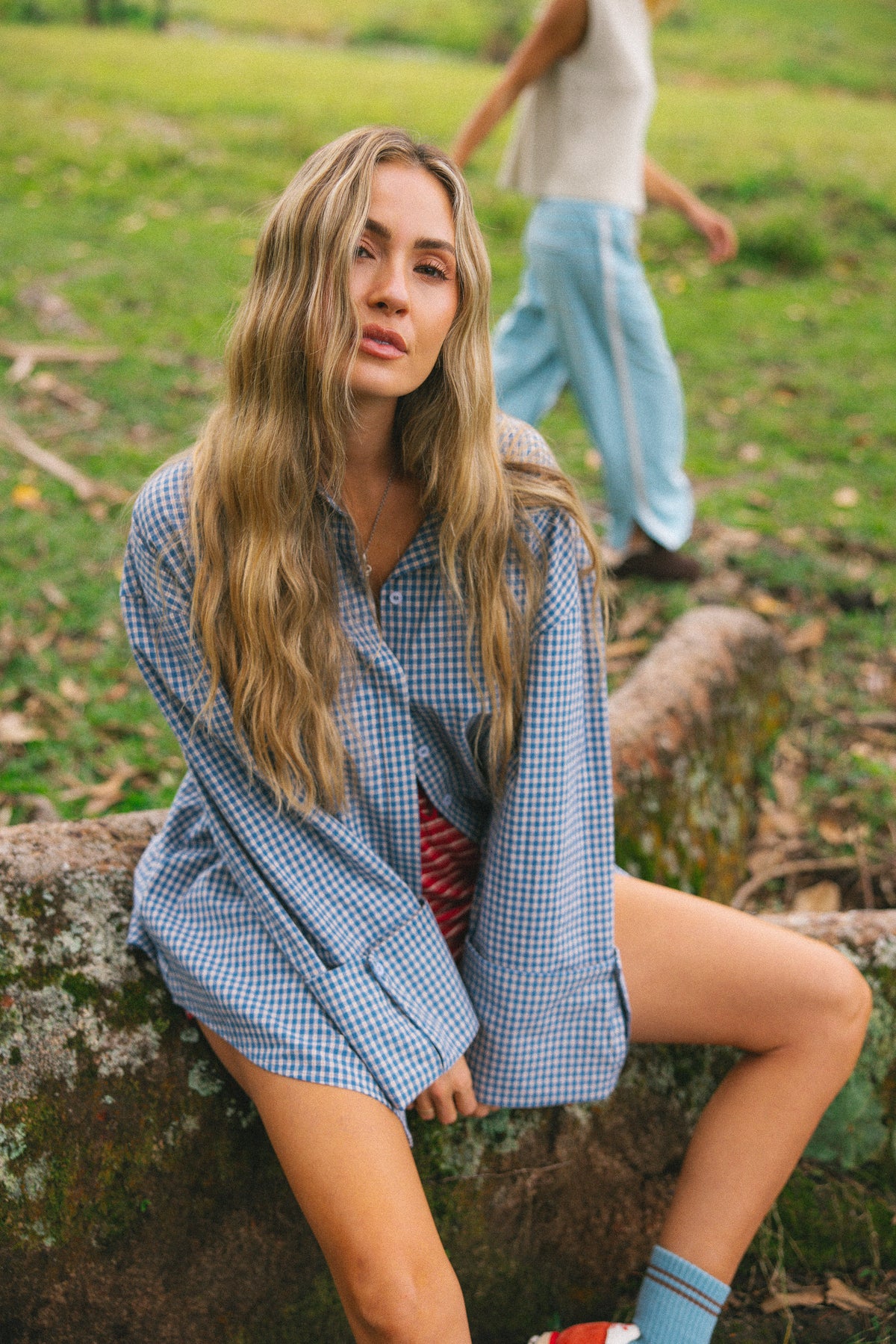 A woman with long blonde hair sits on a mossy log outdoors, wearing an oversized blue and white gingham button-down shirt over a red striped top, paired with light blue socks and red-and-white sneakers. A blurred person walks in the background on a grassy field.