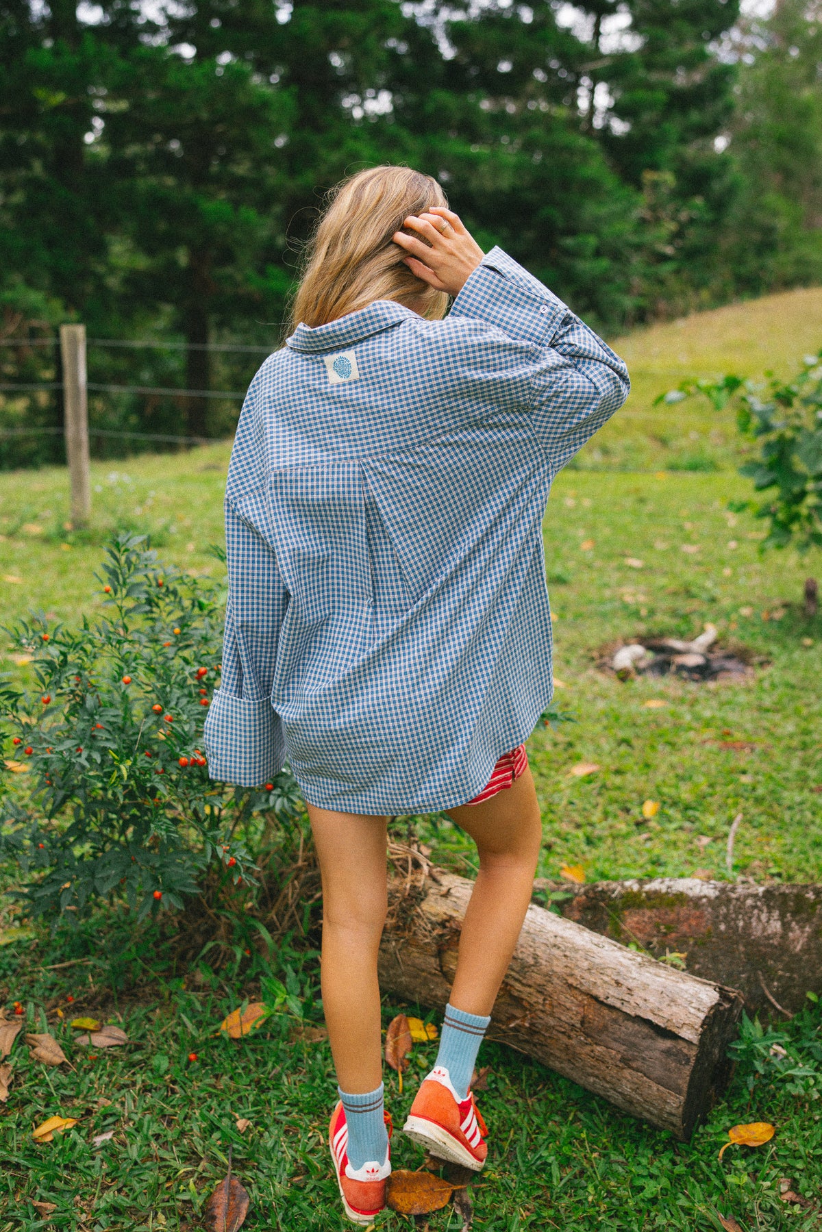 A woman stands outdoors in a grassy yard wearing an oversized light blue and white gingham button-down shirt, red shorts, and orange sneakers.
