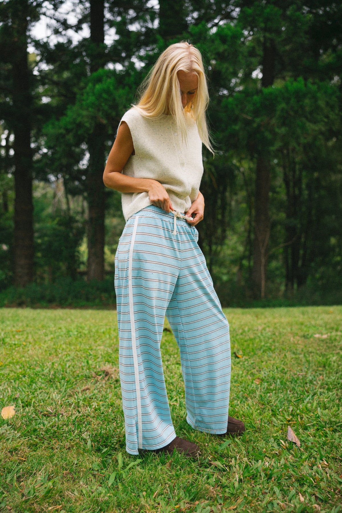 A woman standing on a grassy lawn in front of a forest, wearing light blue and white striped wide-leg pants with a cream sleeveless knit top.