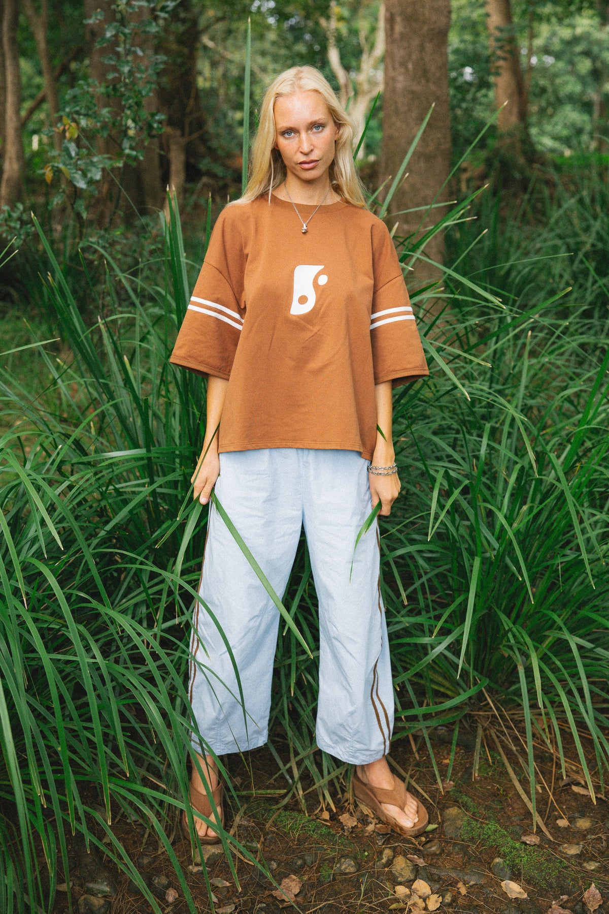 A woman stands in a grassy forest clearing wearing an oversized rust-brown t-shirt with white sleeve stripes and a white abstract logo, paired with light blue cropped pants.