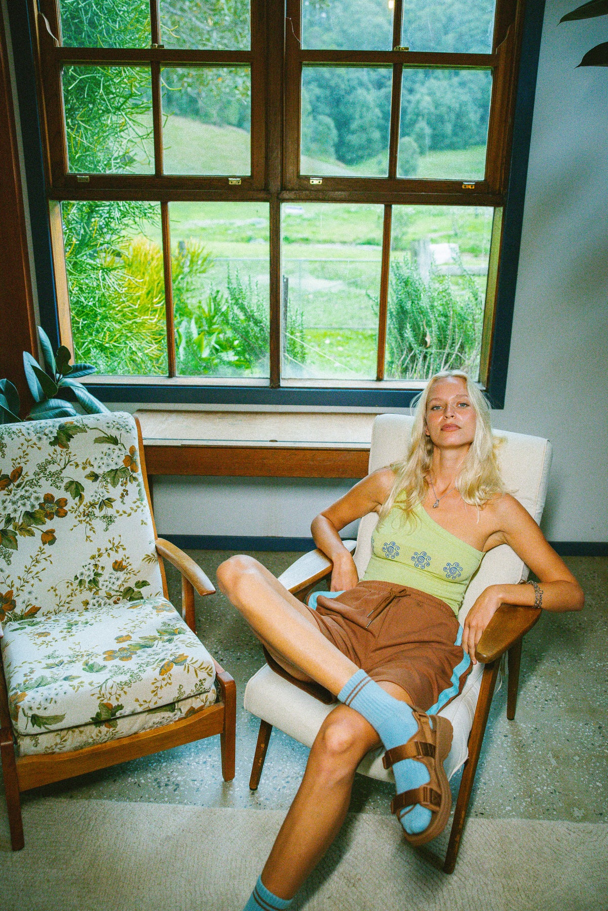 A woman reclines in a white armchair in a bright room with a large window overlooking green fields. She is wearing a light green tank top, brown shorts, and blue socks.