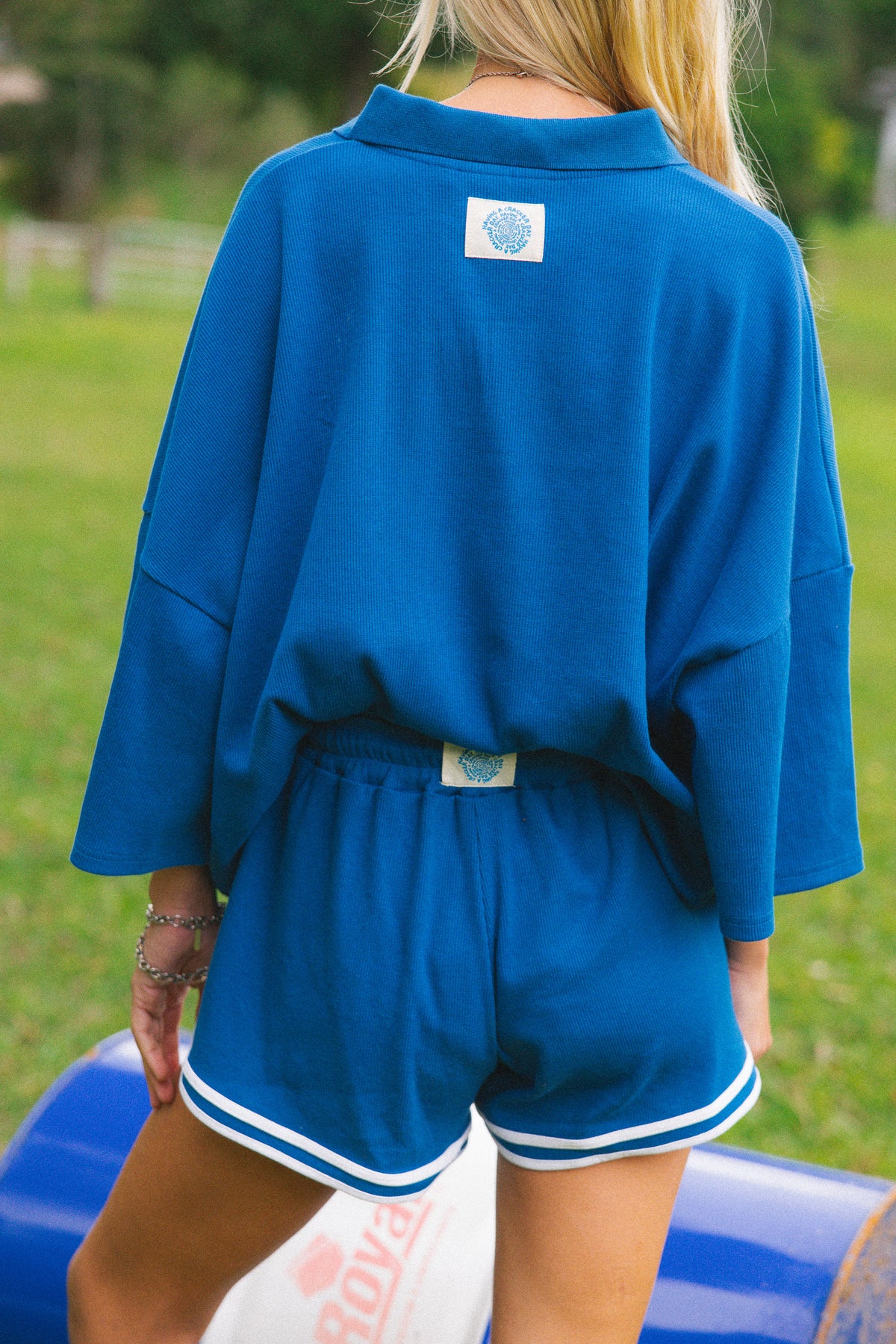 A woman shown from behind wearing a matching royal blue lounge set with a long-sleeve top and shorts featuring white trim. The set has a small white square patch on the back of the collar and waistband.