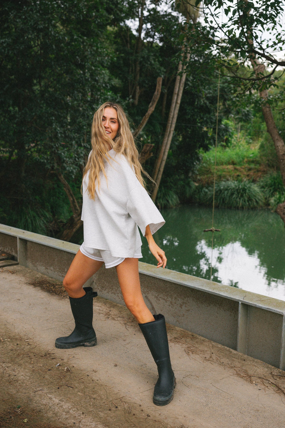 A woman posing outdoors on a dirt path next to a body of water, wearing a grey oversized tunic top, grey shorts, and black rain boots.