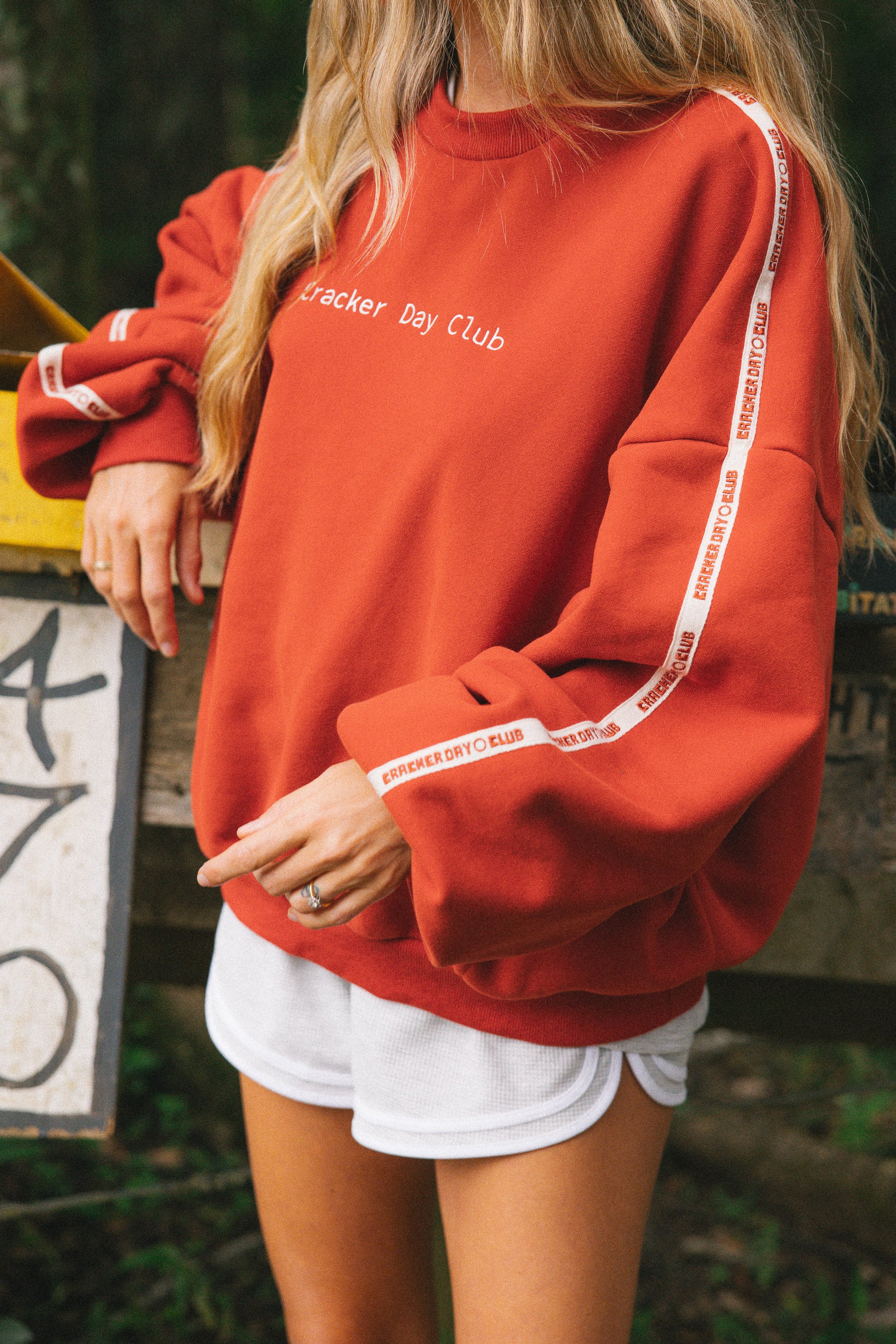 A woman wearing a rust red crewneck sweatshirt with white text reading 'Cracker Day Club' and white tape-style stripes down the sleeves, paired with white shorts, photographed outdoors.
