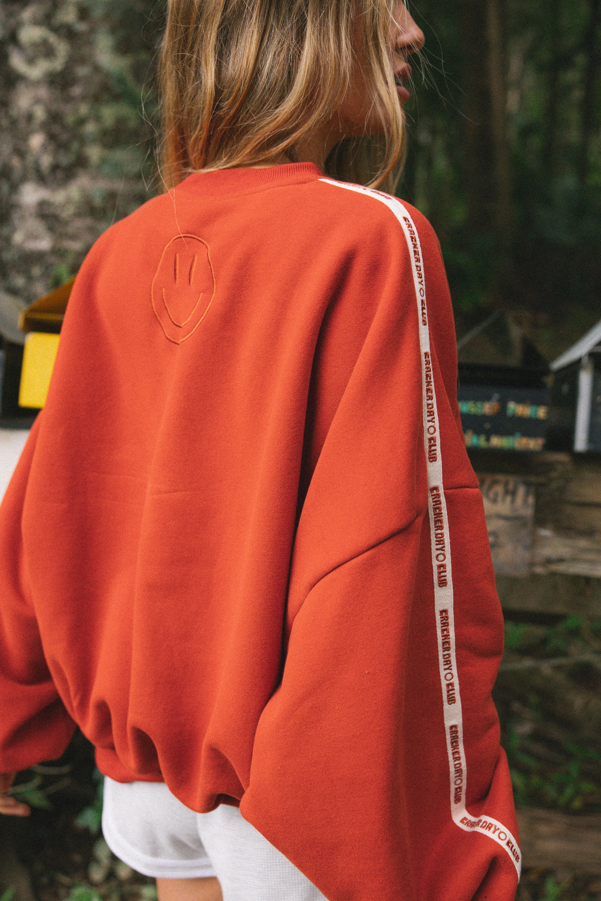 A woman wearing an oversized red sweatshirt with a white tape detail down the sleeve saying cracker day club, photographed outdoors in a forest setting.