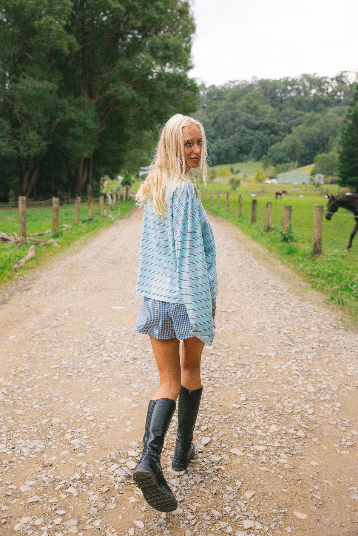 A woman stands on a gravel country road wearing a light blue and white striped long-sleeve shirt, a blue and white gingham mini skirt, and black rain boots, with trees and a field in the background.