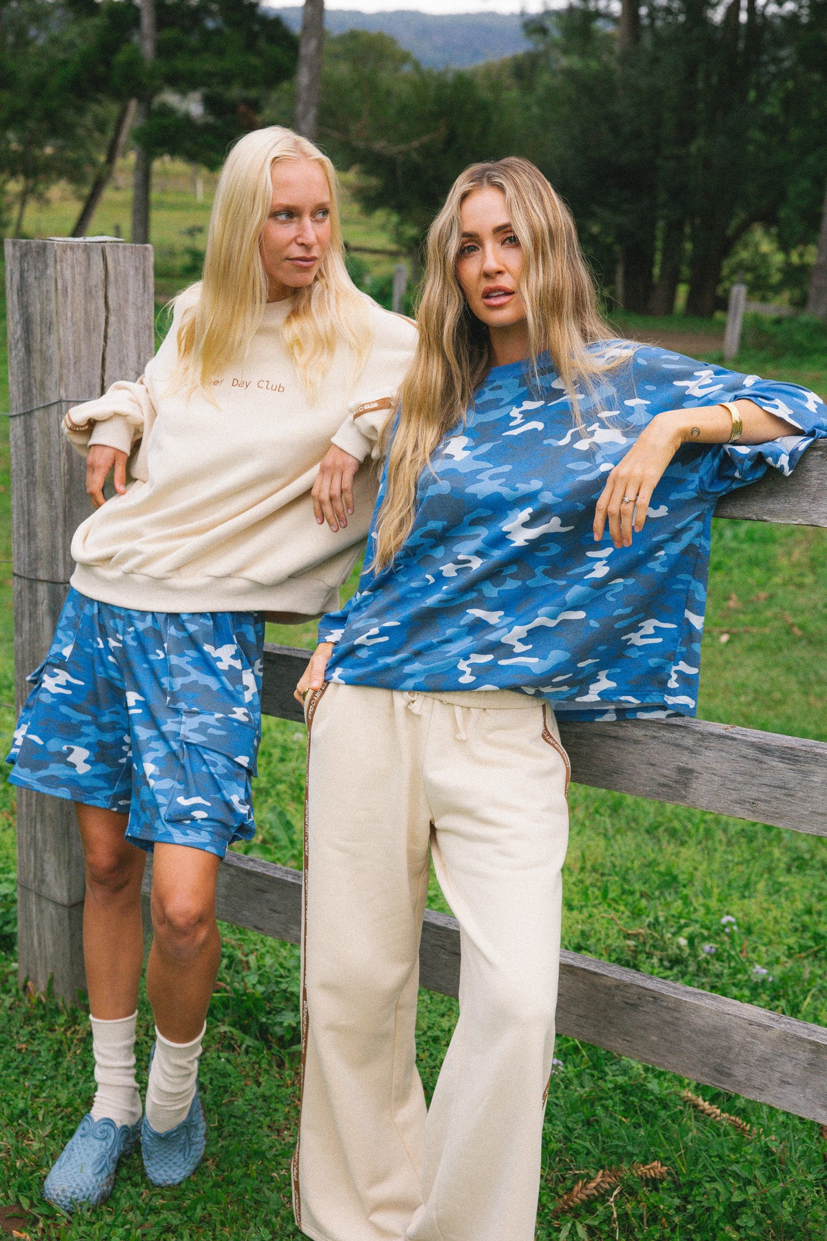 Two women posing outdoors in matching blue and white camouflage lounge sets, with one wearing a cream sweatshirt and blue camo shorts and the other wearing a blue patterned top and cream pants, standing behind a wooden fence in a grassy field.