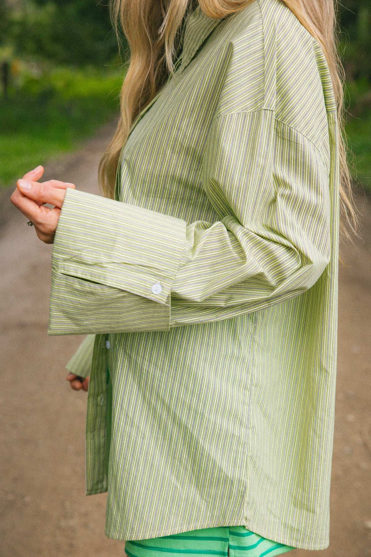 A woman wearing a light sage green and white vertically striped button-up shirt, photographed outdoors on a blurred path with greenery in the background.