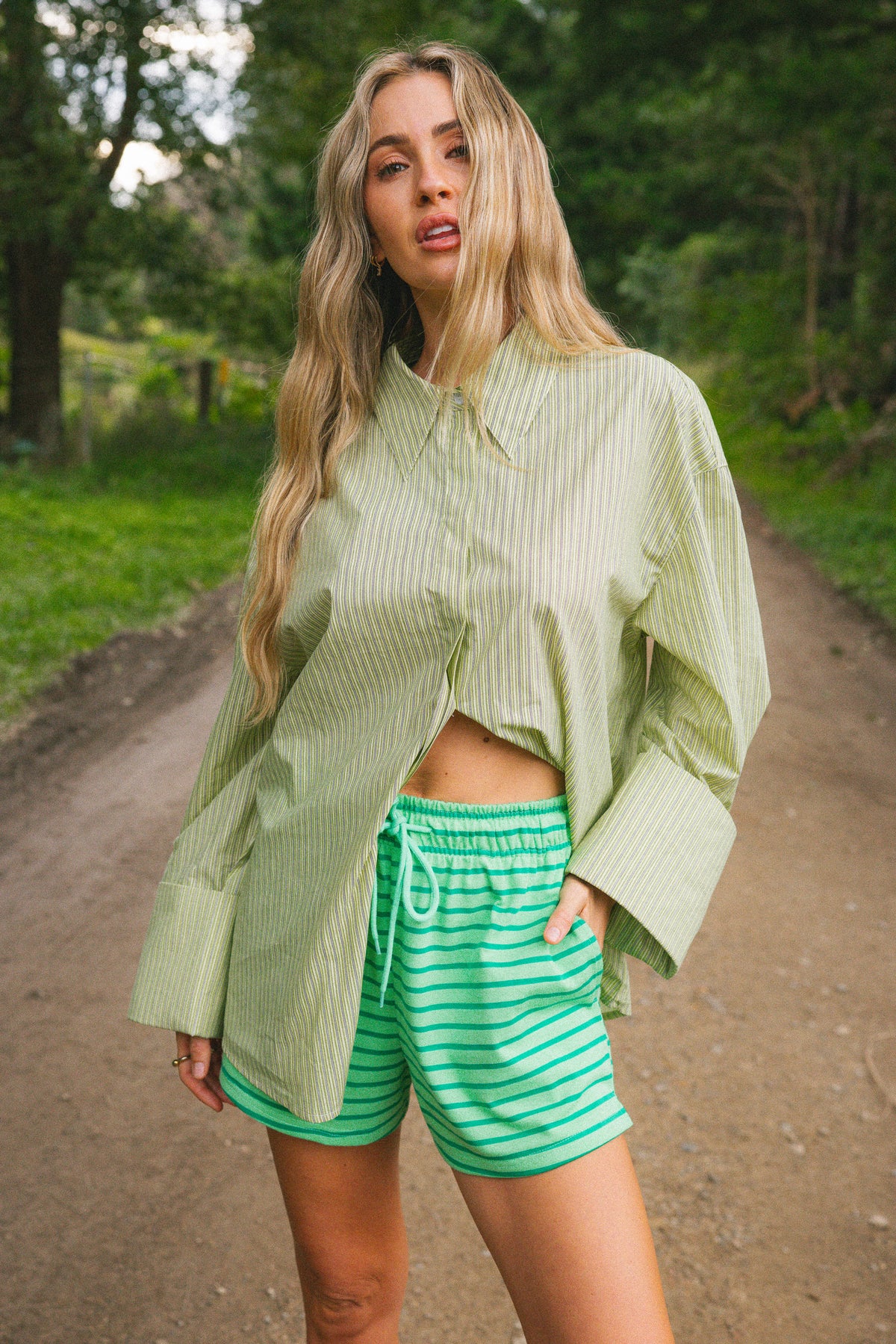 A woman standing on a dirt path in a wooded outdoor setting, wearing a light green vertically striped button-up shirt and green-and-white horizontally striped shorts.