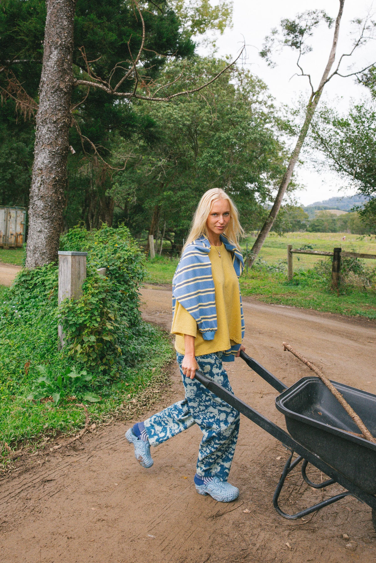 A woman walks along a dirt path outdoors, pushing a black wheelbarrow. She is wearing a yellow top, blue patterned jeans, and a blue striped shawl.