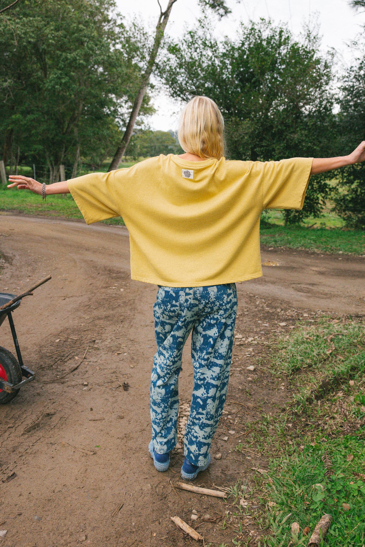 A person standing outdoors on a dirt path with arms outstretched, wearing a loose bright yellow oversized T-shirt and blue-and-white patterned pants.