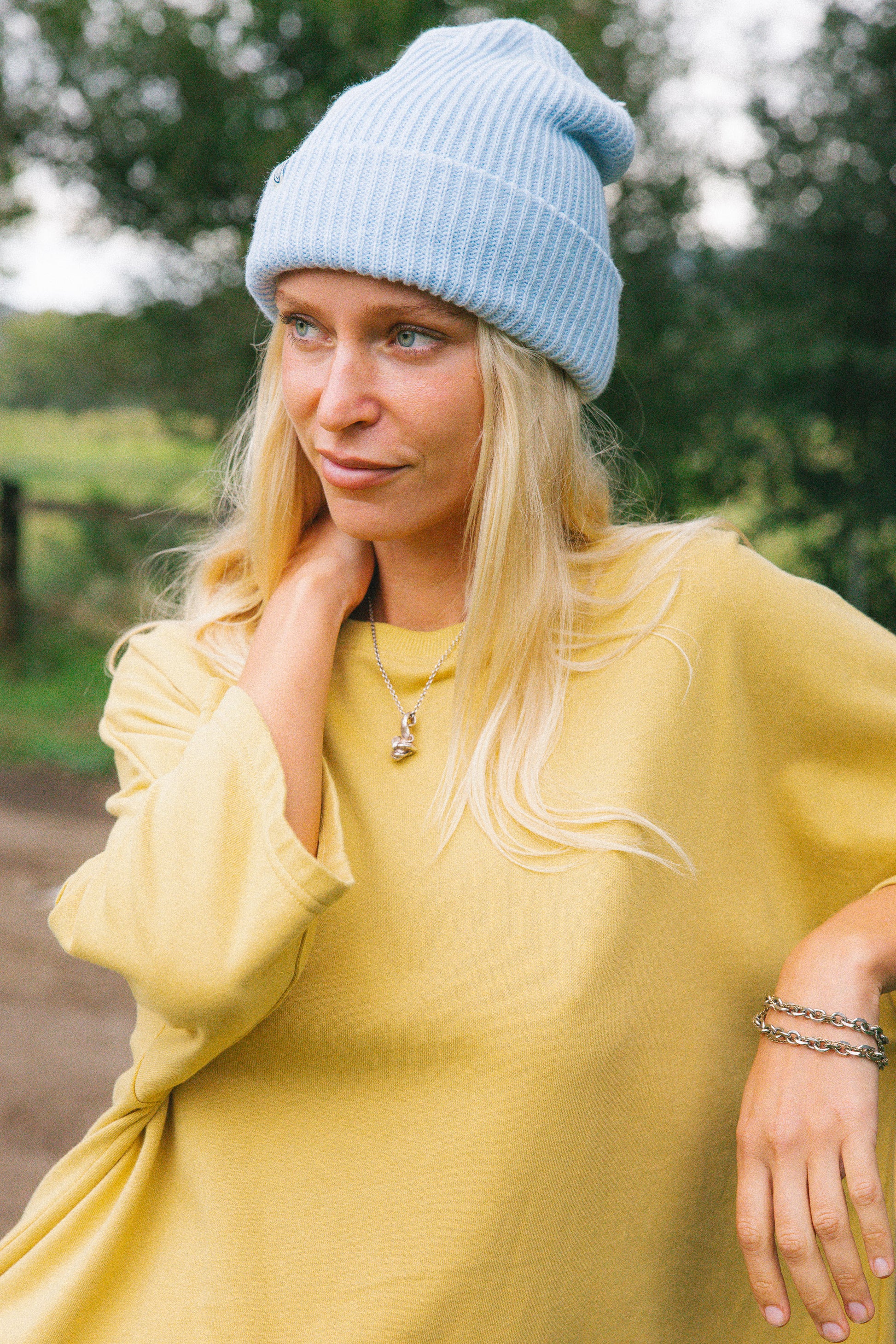 A woman wearing a light blue ribbed knit beanie and a loose mustard yellow long-sleeve top, photographed outdoors with a blurred green background.