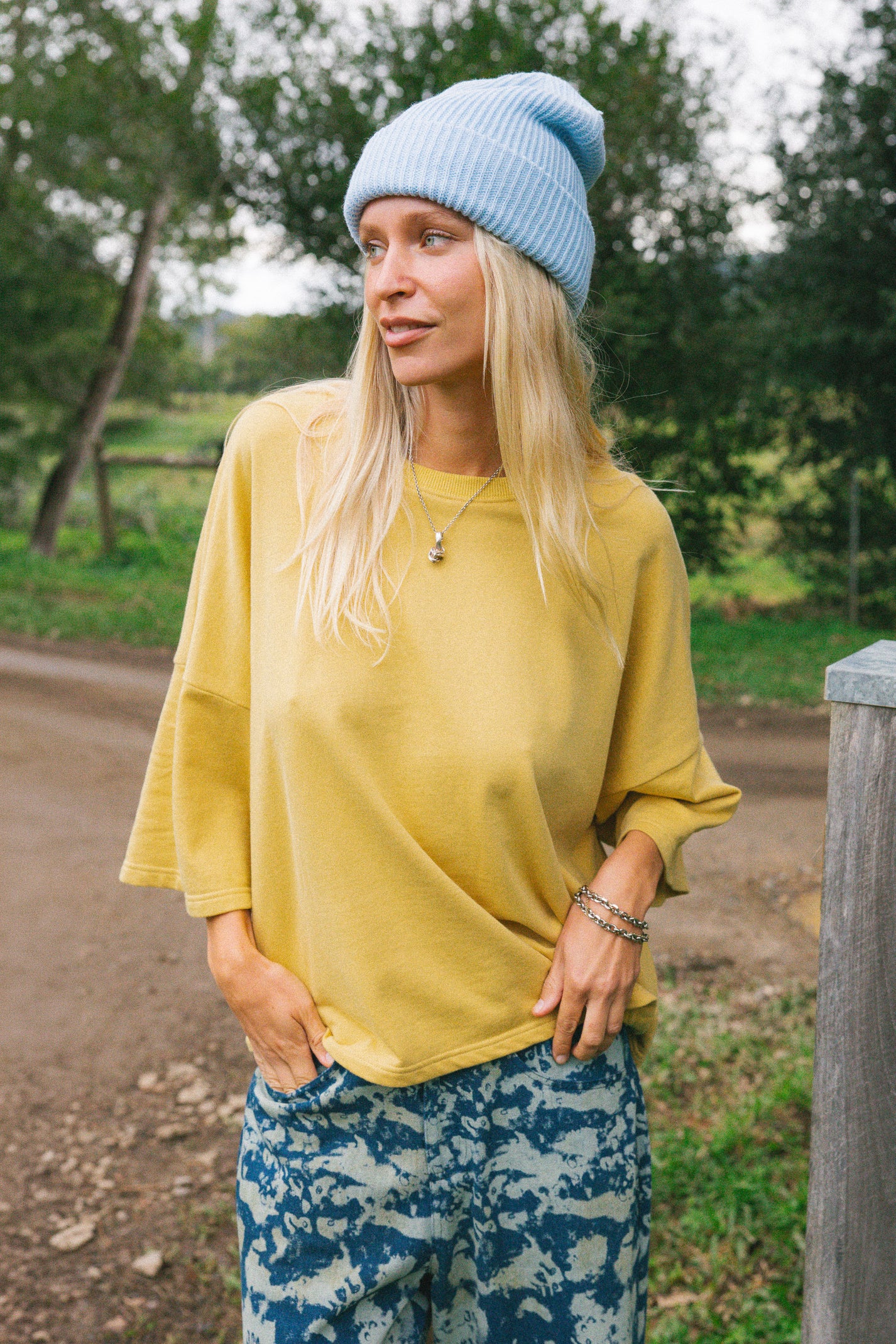 A woman wearing a light blue knit beanie, a mustard yellow oversized long-sleeve top, and blue-and-white patterned pants, standing outdoors on a dirt path with trees in the background.