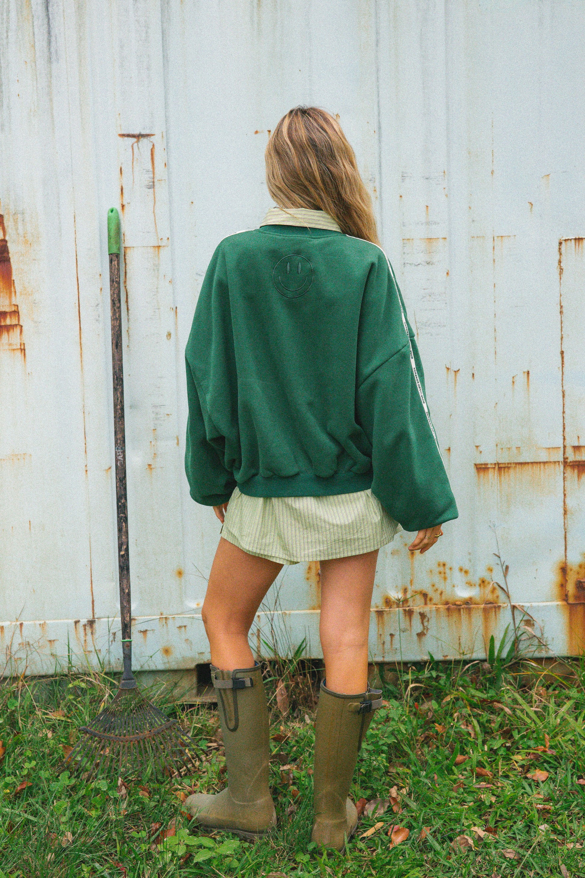A woman viewed from behind wearing a dark green crewneck sweatshirt and olive green rain boots, standing in front of a weathered white wooden fence.
