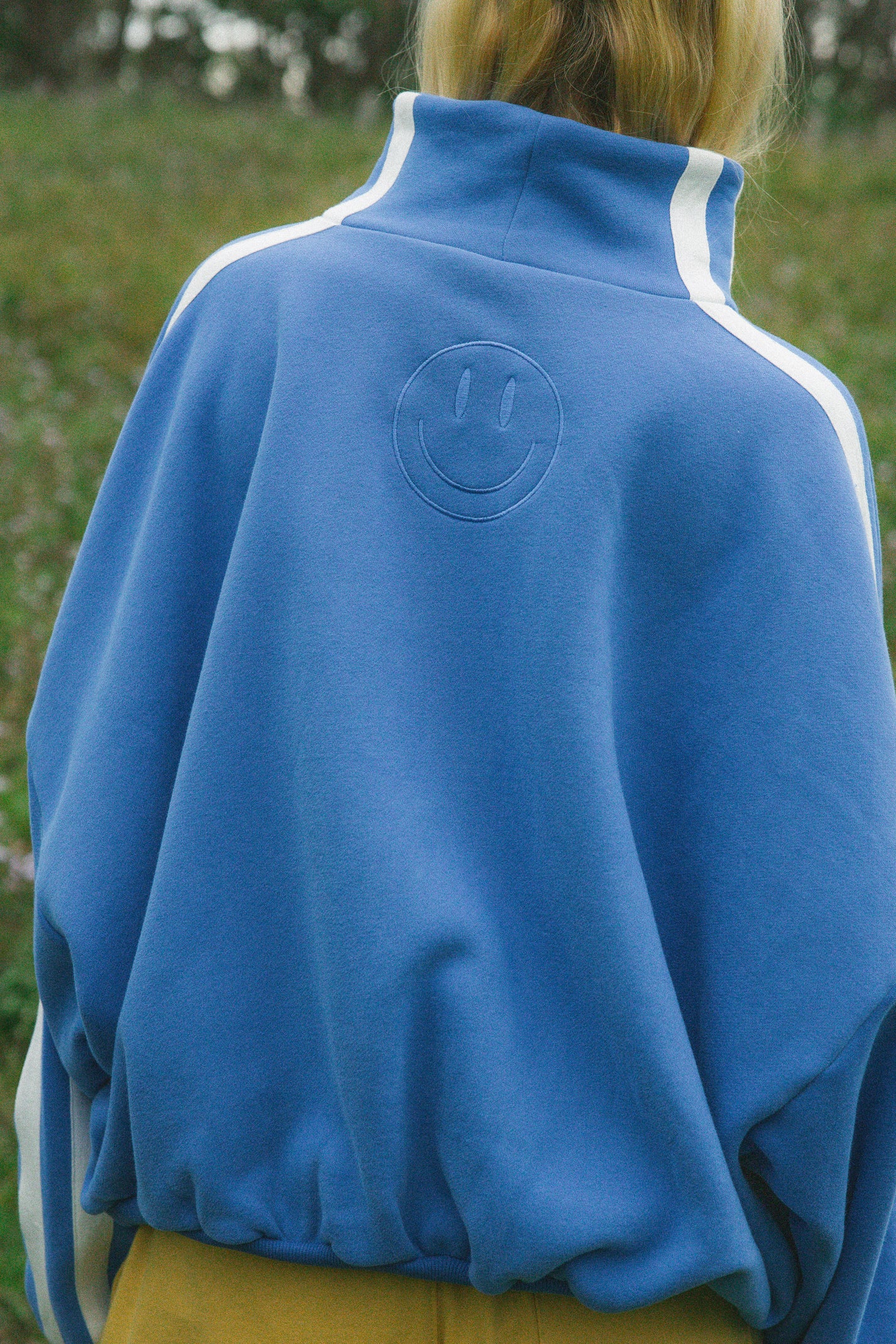 A person viewed from behind wearing a blue sweatshirt with white shoulder stripes and an embossed smiley face on the back, photographed outdoors on a grassy background.