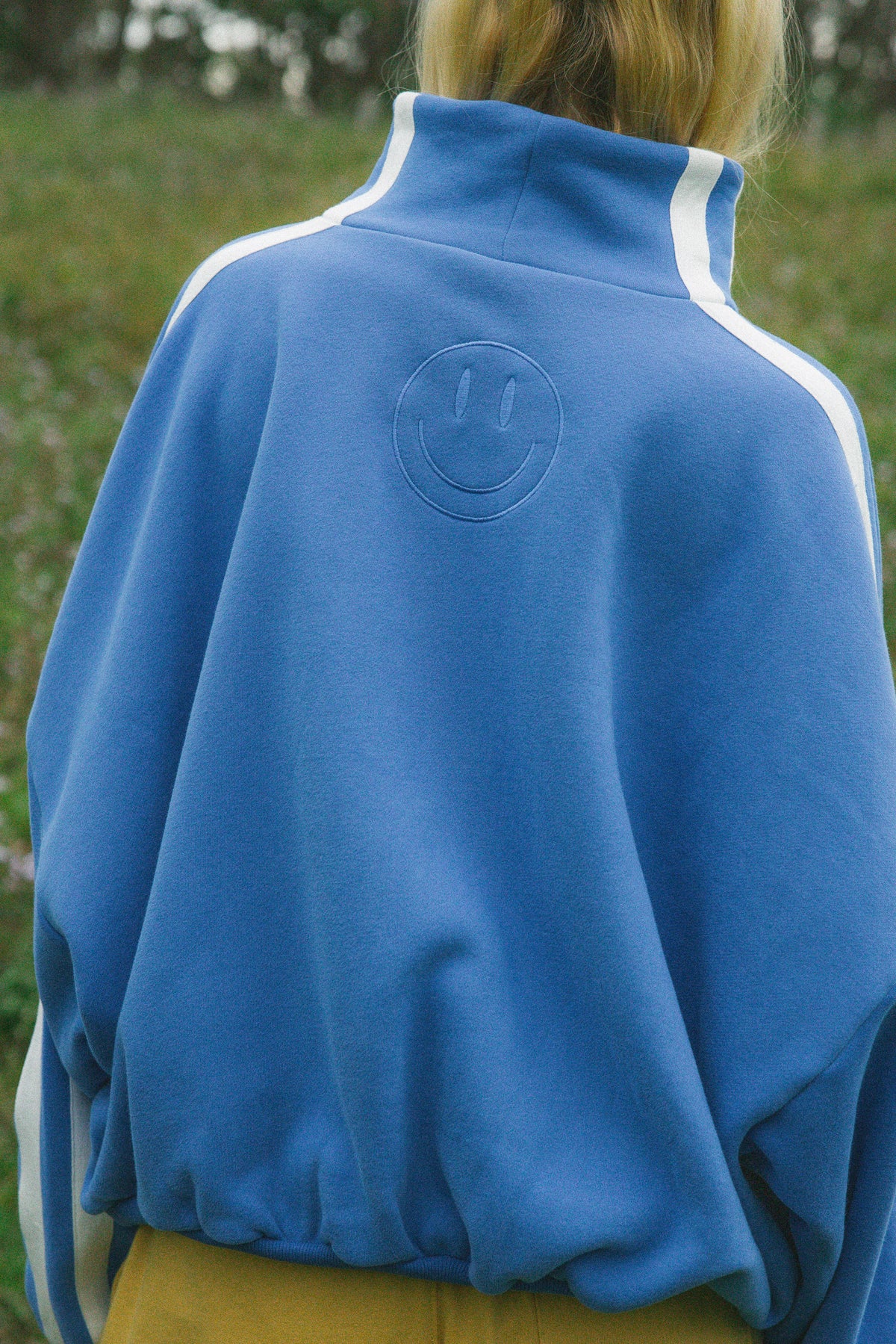 A person viewed from behind wearing a blue sweatshirt with white shoulder stripes and an embossed smiley face on the back, photographed outdoors on a grassy background.