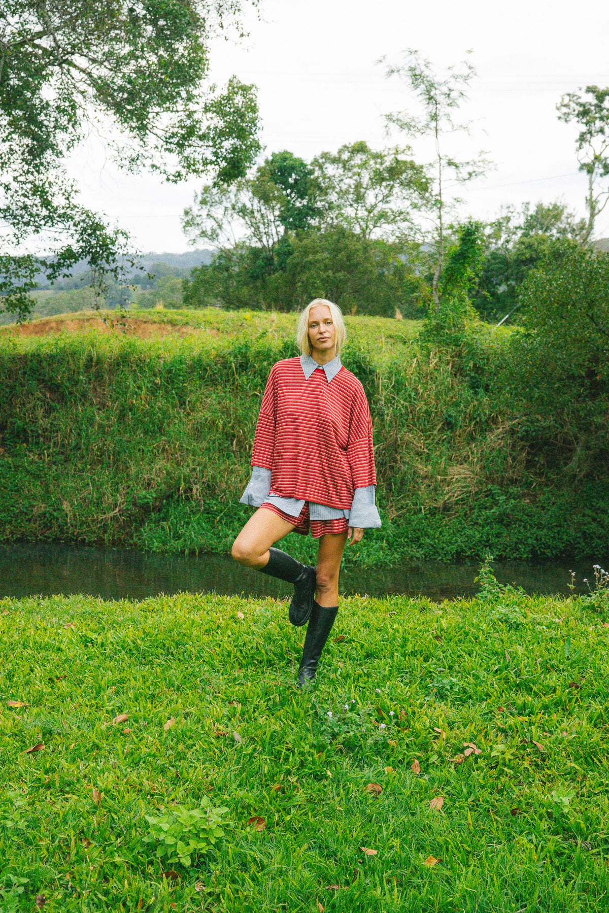 A woman stands in a grassy outdoor setting near a small stream, wearing a red-and-white striped poncho-style top with light blue cuffs, a matching striped skirt, and black rain boots.