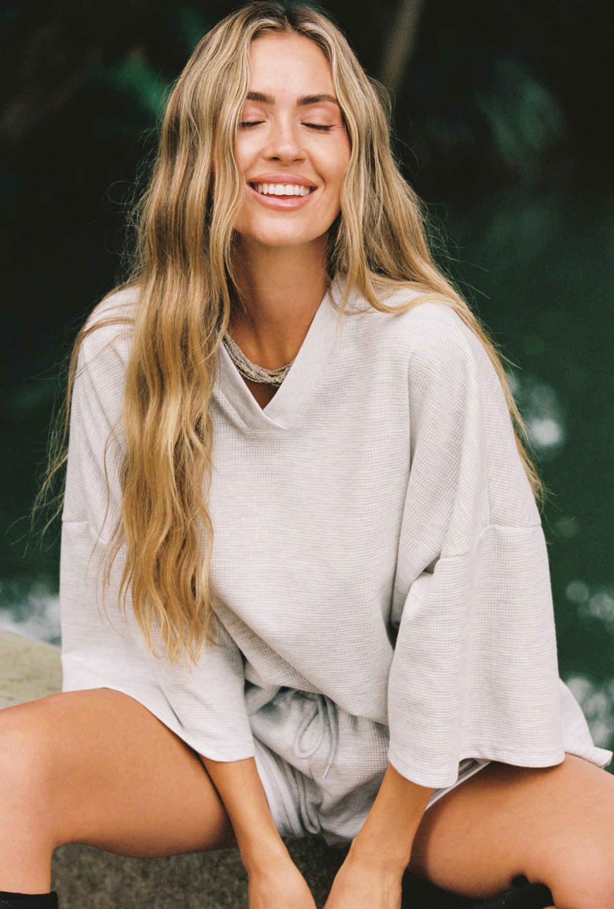 A woman with long blonde hair wearing a light gray oversized waffle top and matching shorts, sitting outdoors against a dark green blurred background.