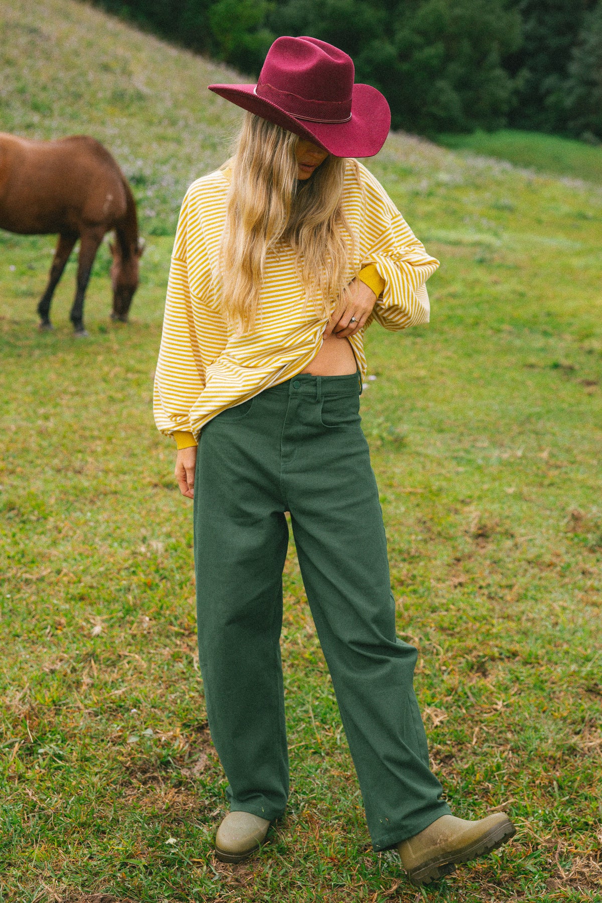 A woman standing on a grassy hill wearing a yellow striped long-sleeve shirt, dark green wide-leg pants, a burgundy wide-brim hat, and tan boots, with a horse visible in the background.