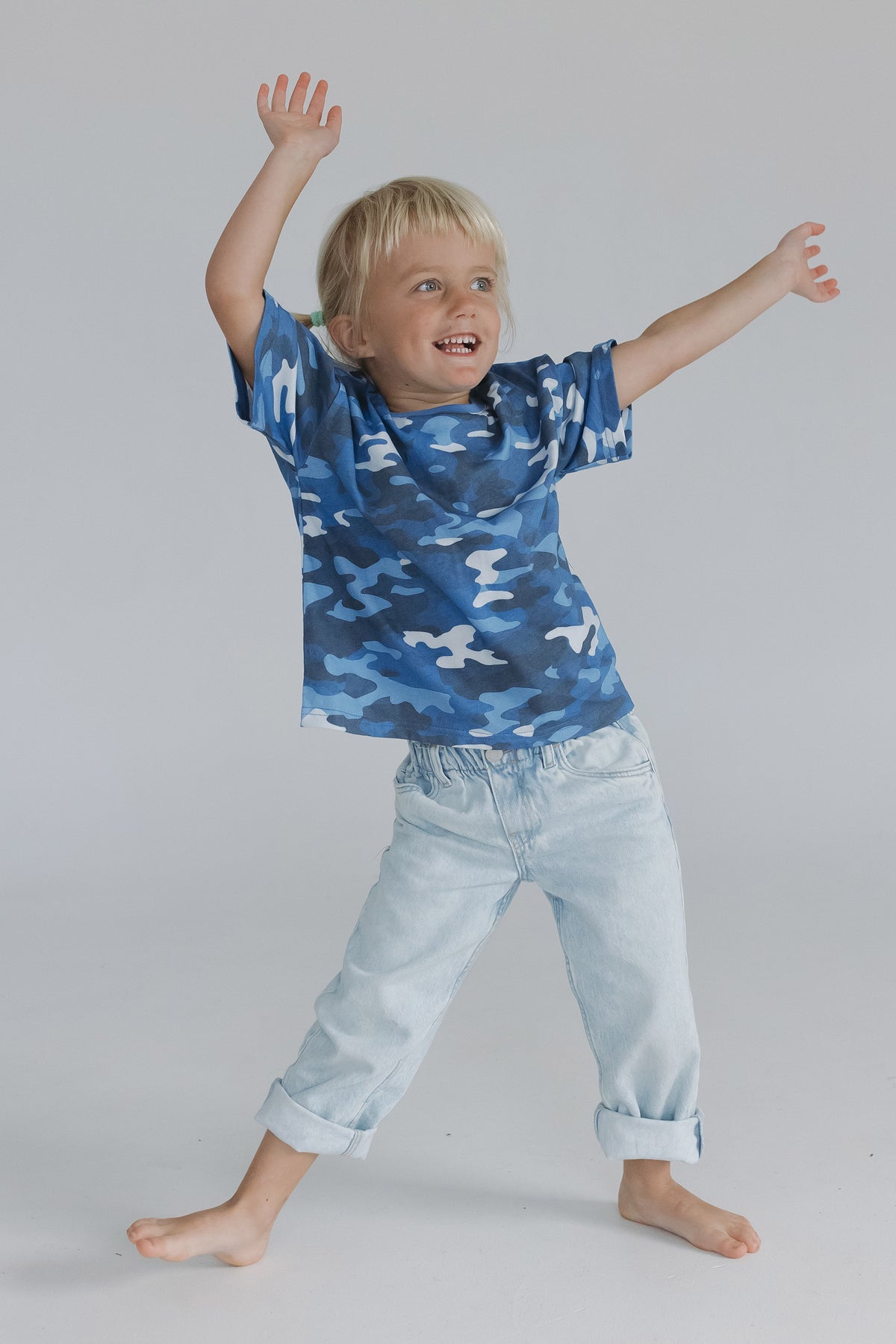 A young blonde girl wearing a blue camouflage t-shirt and light wash jeans, posing with his arms outstretched on a seamless light gray studio background.