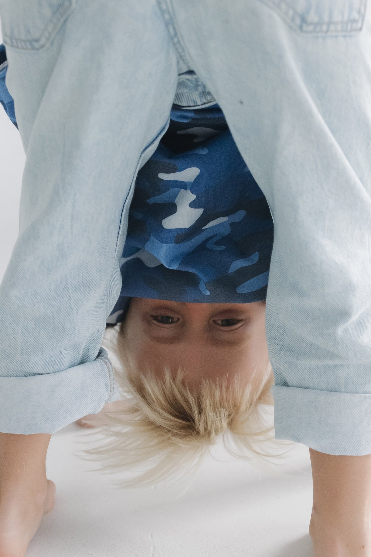 A child wearing a blue and white camouflage t-shirt under a light-wash denim shirt, performing a handstand on a white floor.