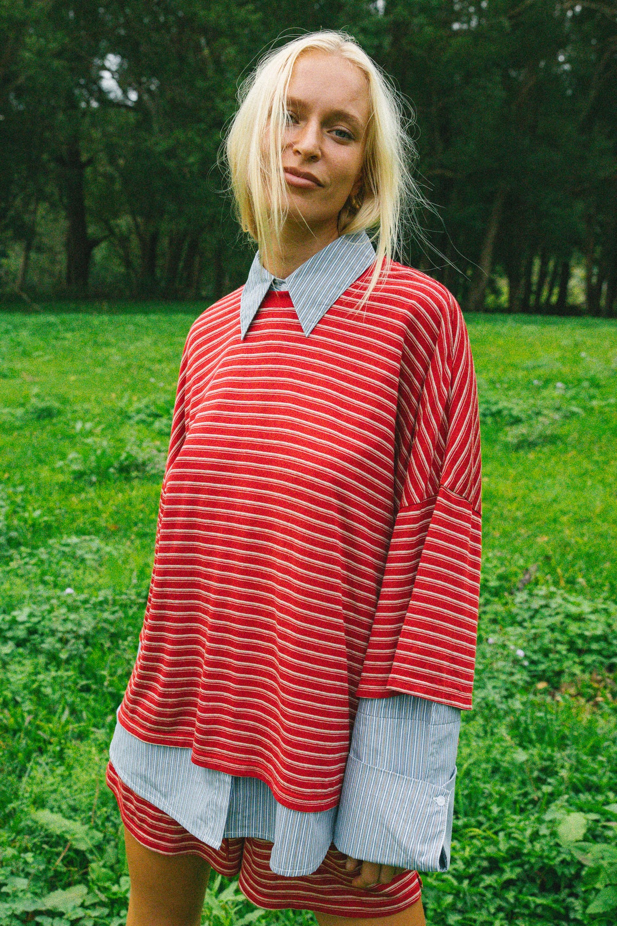 A woman standing outdoors in a grassy park wearing a red and white striped oversized tunic top with light blue and white striped collars and cuffs, paired with matching striped shorts.