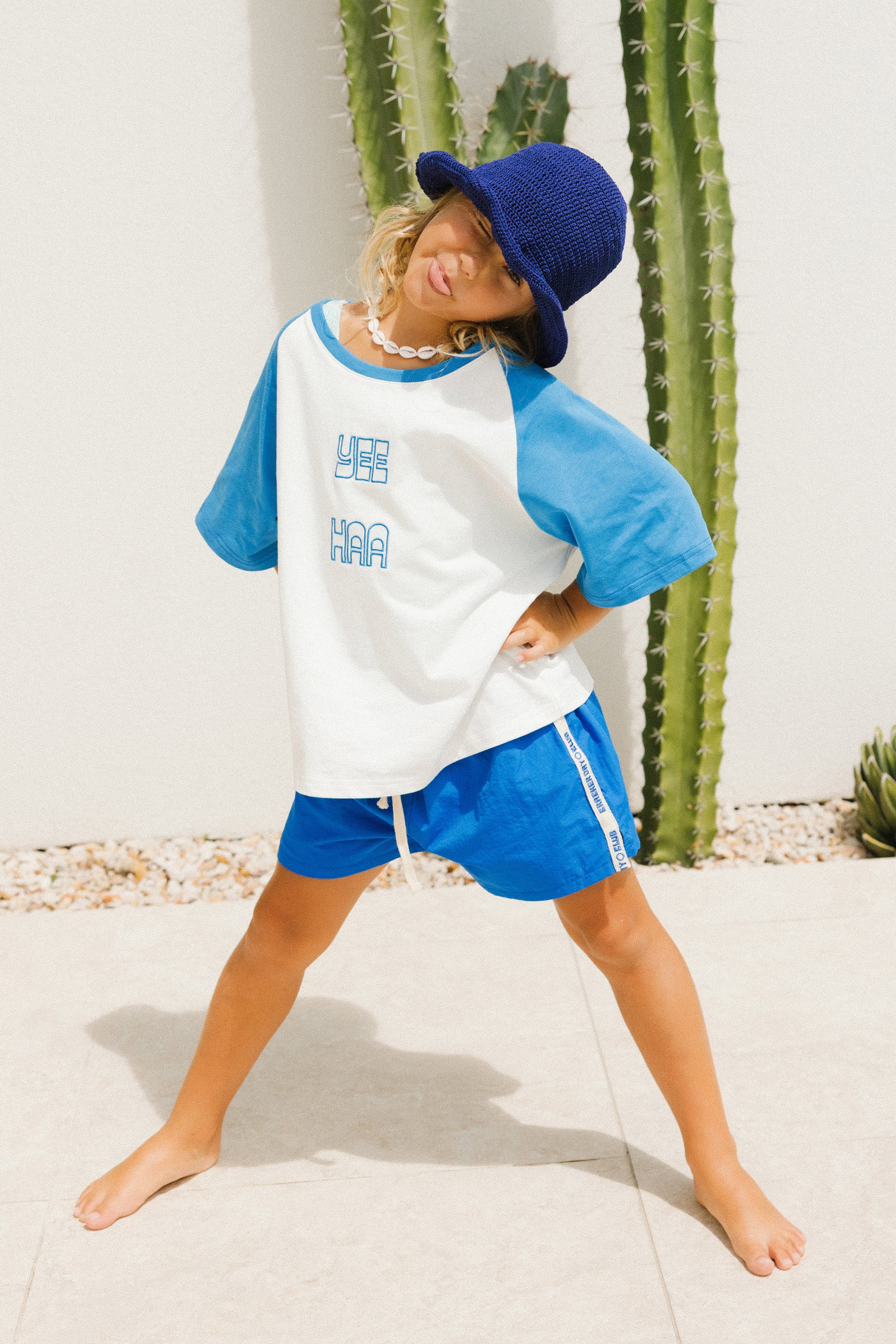 Child wearing a blue and white baseball shirt with yee haa on the front and shorts with a blue cap, standing in front of cacti.