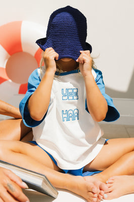 Child wearing a white t-shirt with blue text and blue boardshorts, sitting on a white surface.