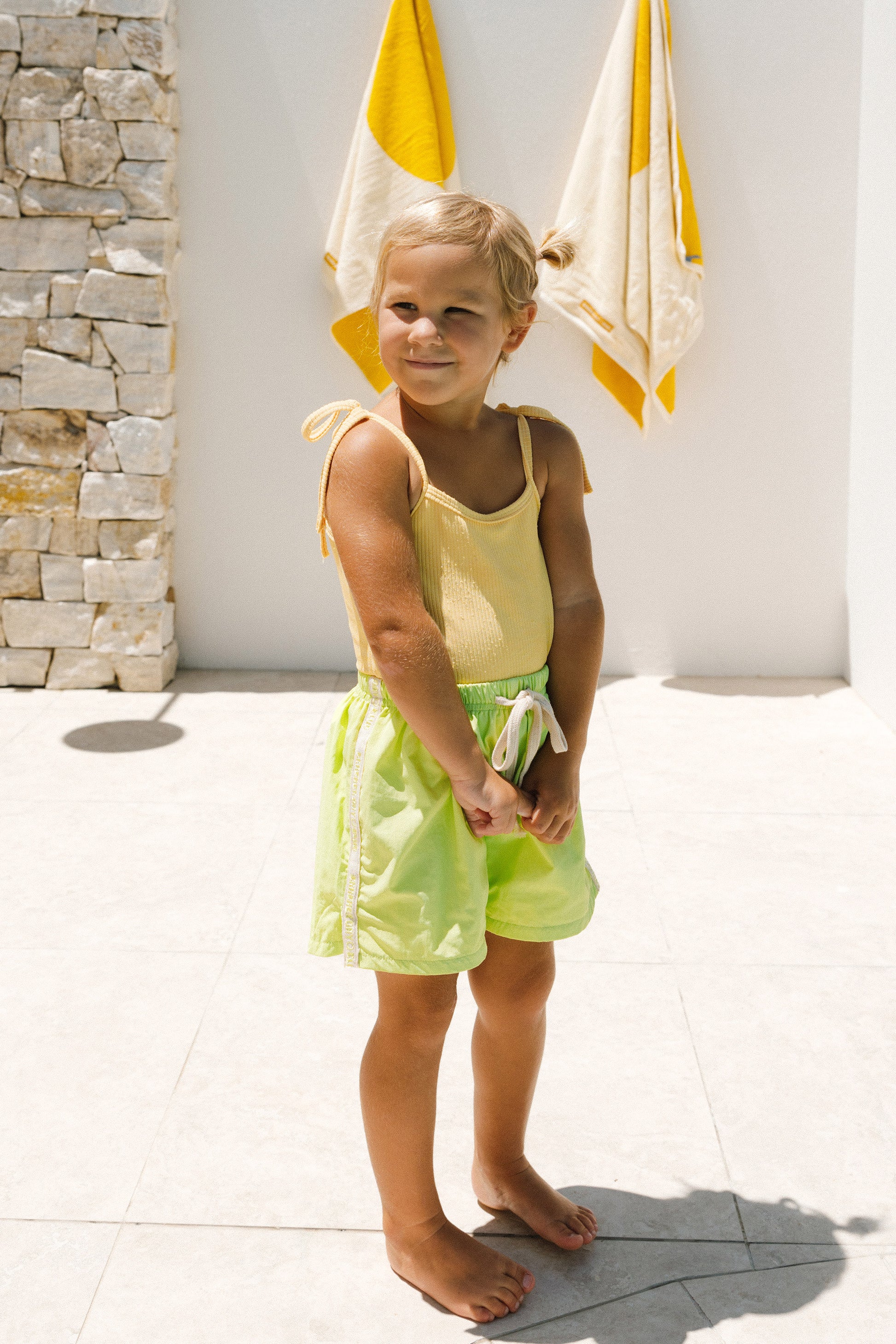 Young girl in yellow top and green cracker day boardshorts standing in front of a stone wall and white wall with yellow towels.