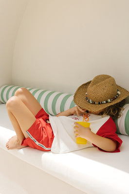 Child wearing a straw hat and red shorts lying on a white couch with green striped cushions.