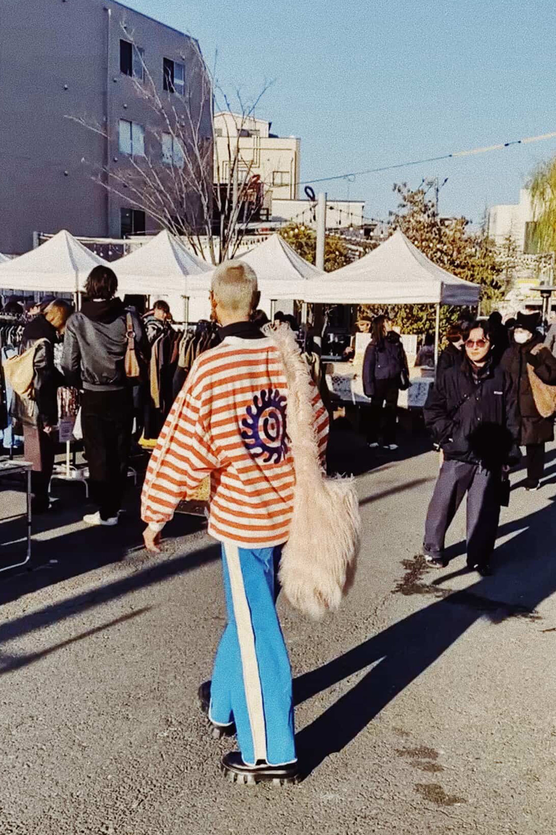 Person wearing a striped jacket and blue pants walking on a street with tents and people in the background.
