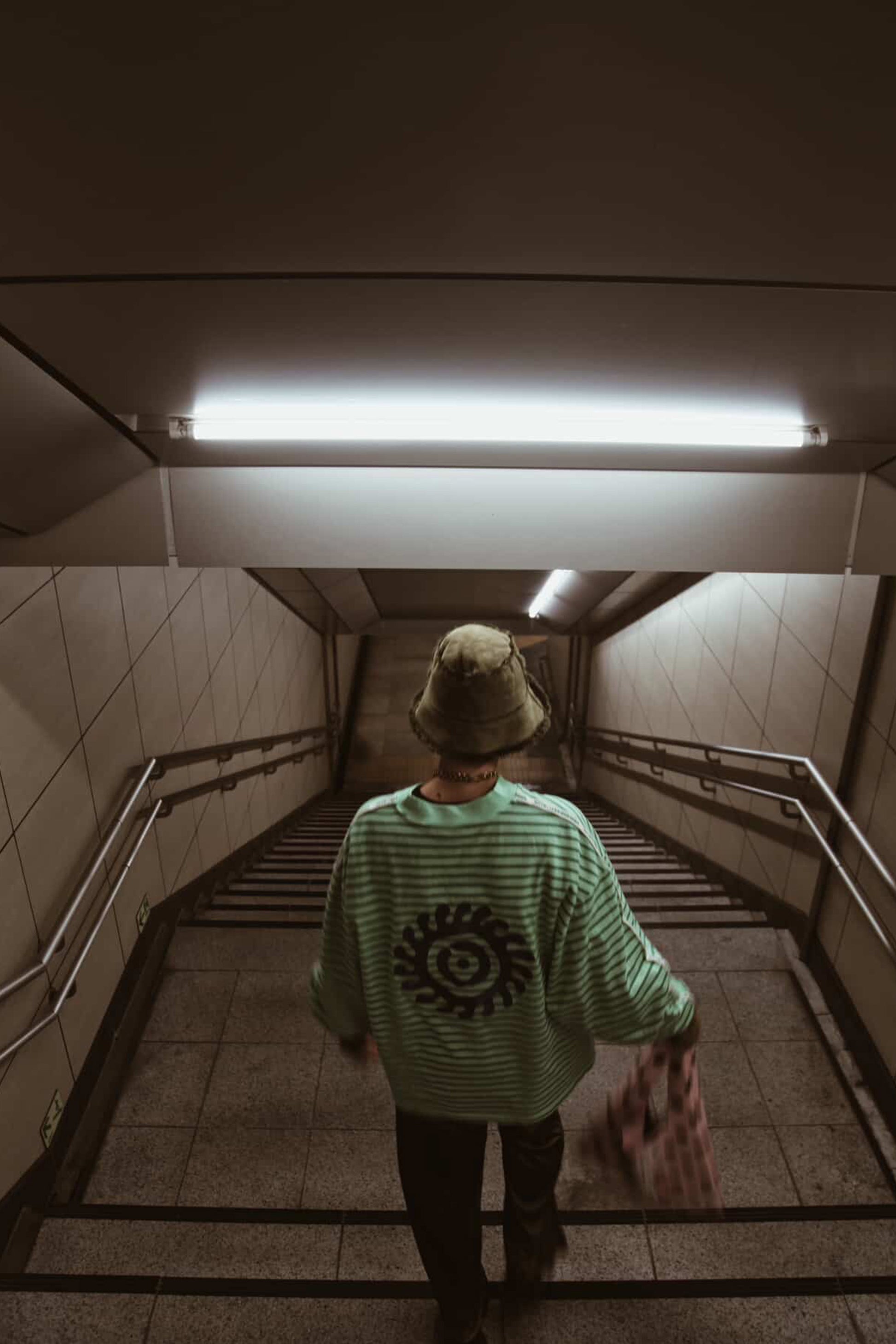 Person walking down a staircase in a dimly lit hallway with a green sweater and bucket hat.
