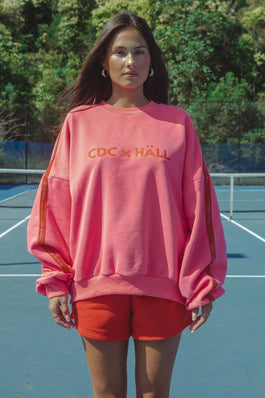 Woman wearing a pink sweatshirt with cracker day club x hall text on a tennis court
